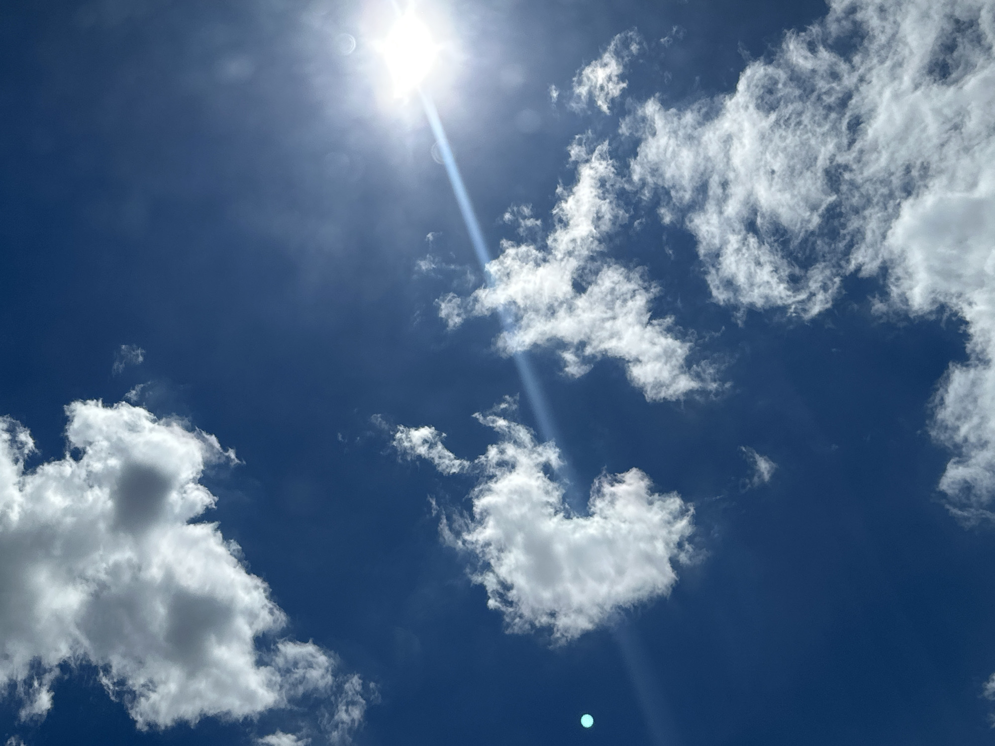 The image displays a bright sun shining through a deep blue sky dotted with scattered white clouds. The sun is positioned in the upper left quadrant of the frame, emitting a strong glare and visible light rays. Some clouds are thin and wispy, while others are more substantial and rounded. The overall impression is of a clear, sunny day. There are no people, structures, or specific geographical features visible that would indicate a location within a city or any specific environment. The image is a straightforward depiction of the sky on a sunny day.