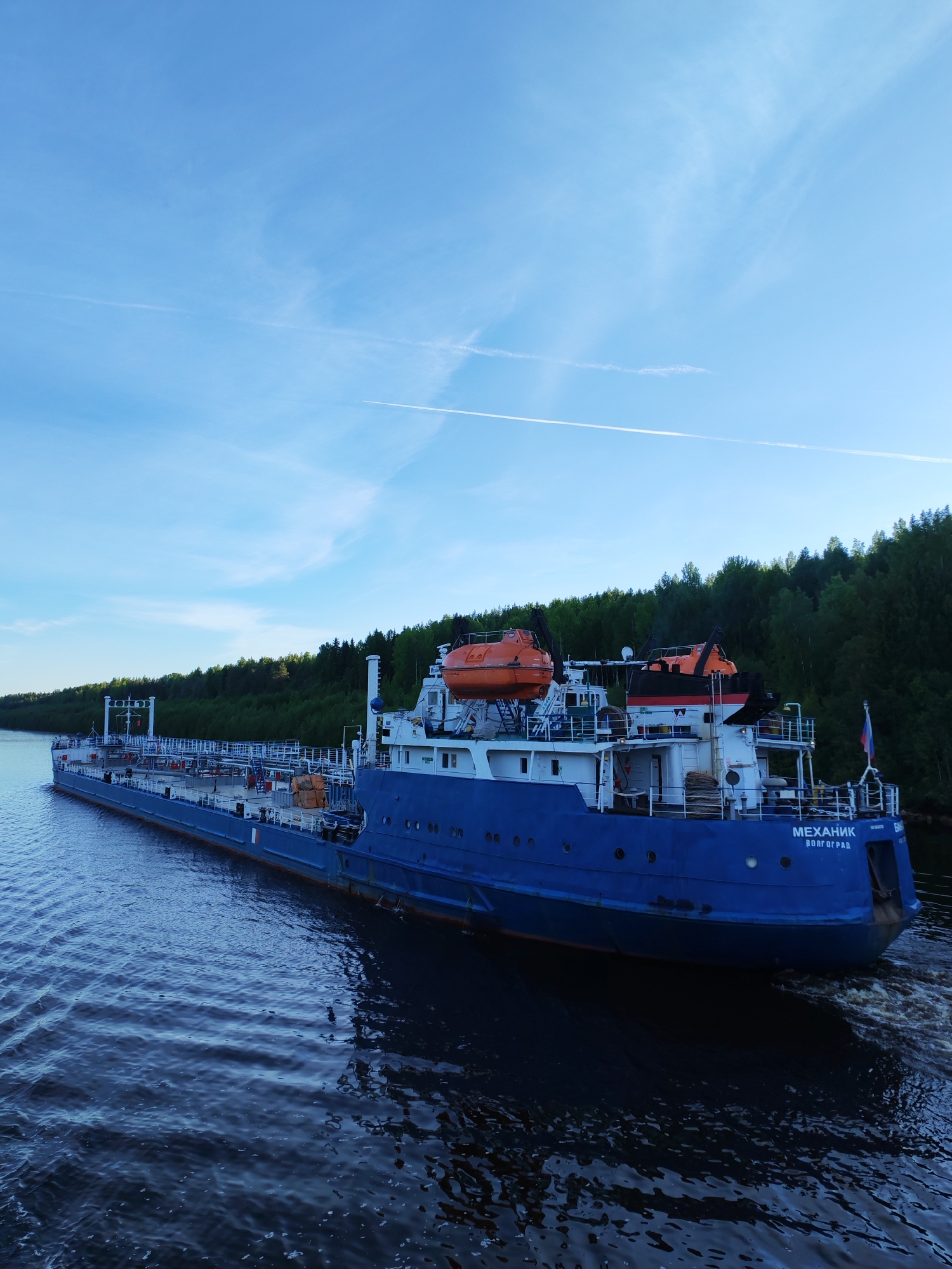 A large, dark blue tanker ship or barge is the central subject, navigating a calm body of water. The vessel is equipped with a white superstructure and two prominent orange enclosed lifeboats positioned on davits towards its stern. Numerous portholes are visible along its hull and superstructure, and railings line the decks. A Russian flag flies from the stern.
The ship is actively moving, as indicated by the churning wake trailing behind it. The scene is set on what appears to be a river or canal, with a dense, verdant forest forming the backdrop along the far bank. The sky above is a clear, bright blue, featuring faint wispy clouds and at least two distinct white airplane contrails stretching across it.
The overall impression is one of peaceful transit during daylight hours, likely mid-morning or late afternoon, given the clear conditions. The text "МЕХАНИК" (MEKHANIK) is clearly visible in white capital letters on the ship's stern, with "ВОЛГОГРАД" (VOLGOGRAD) written underneath, indicating its name and home port. The location context provided places this scene in or near Старое Петровское, Russia.