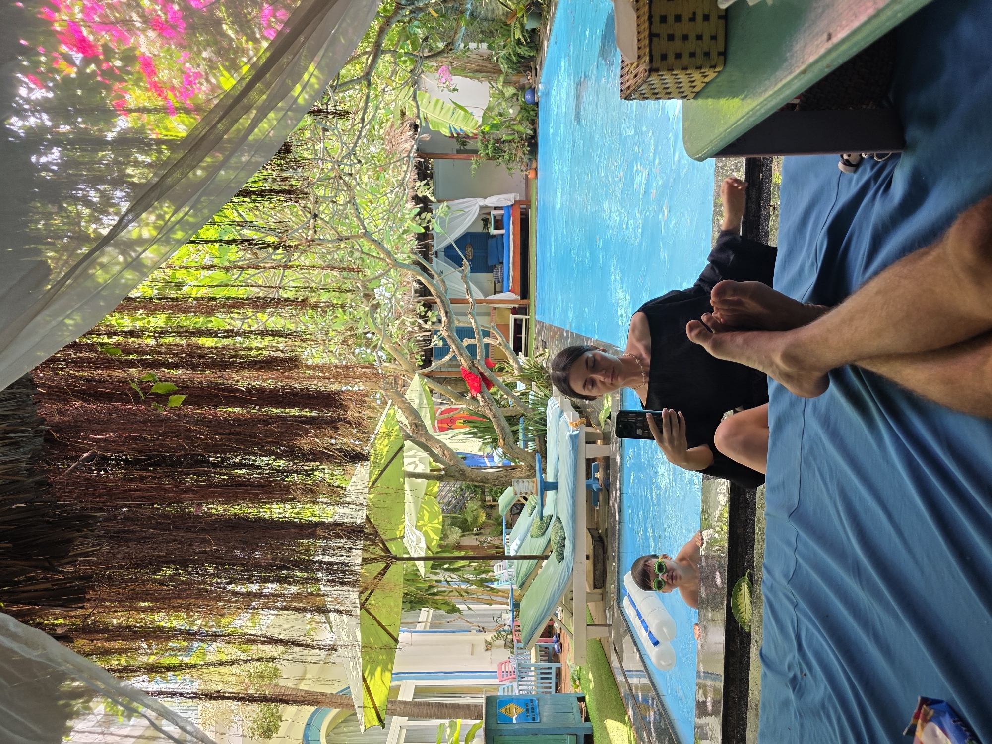 The image captures a scene at the Sailing Club Mui Ne in Mũi Né, Vietnam. A woman is seated, holding a smartphone, seemingly taking a picture or using it near a bright blue swimming pool. A child wearing green sunglasses floats on an inflatable in the pool. Part of a person's leg and foot is visible in the foreground, possibly belonging to someone on a lounger. The surrounding area includes trees with exposed roots hanging down, sun loungers with green cushions, and colorful buildings typical of the resort town. The scene is captured outdoors, under daylight conditions, with a hint of dappled light filtering through the foliage.