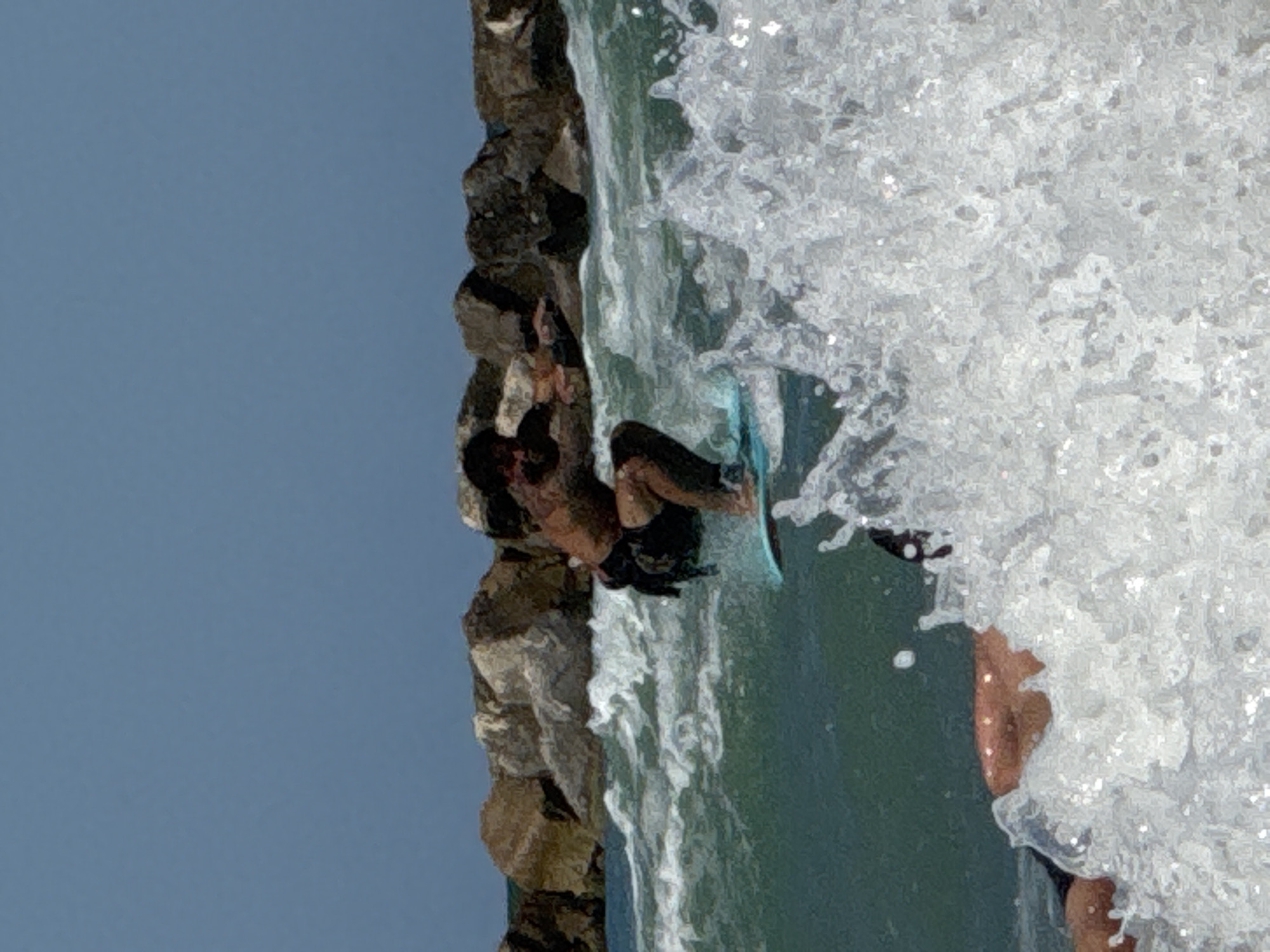 A person is bodyboarding in the ocean near a rock breakwater on a sunny day. The person is wearing dark shorts and has tattoos on their back and arms. The water is a clear turquoise color and the waves are breaking. In the foreground, a large wave is crashing, obscuring the lower part of the image. The background shows a rough breakwater made of large rocks. The sky is a clear blue. The location is likely the Adriatic Sea, as San Benedetto del Tronto is situated on the coast of Italy.