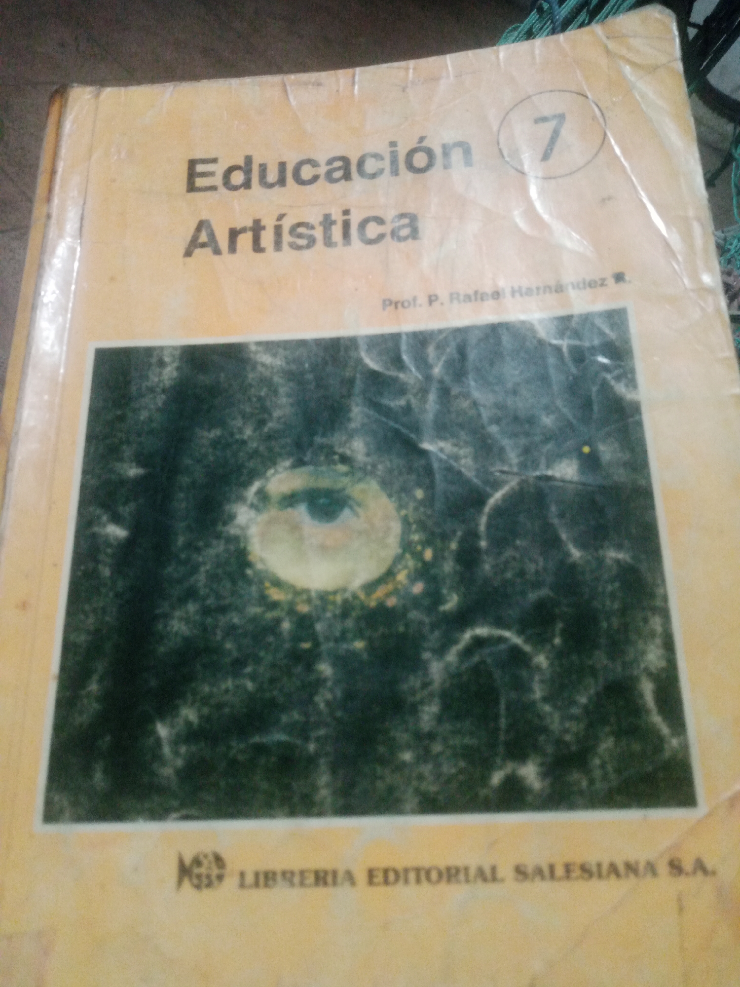 The image shows the cover of a book titled "Educación Artística 7". The cover is a light yellow color with black text. The title "Educación Artística" is in large, bold letters, with "Educación" above "Artística". To the right of the title, there is a circled number "7". Below the title, there is smaller text that reads "Prof. P. Rafael Hernández". The bottom of the cover features an image of an eye against a dark, textured background, with a logo and the text "LIBRERIA EDITORIAL SALESIANA S.A." printed below it. The book appears to be in a somewhat worn condition, with some creasing and discoloration on the cover. The lighting suggests the photograph was taken indoors, likely with natural light. The book's origin or current location in Santa Ana, Venezuela, cannot be determined from the image.