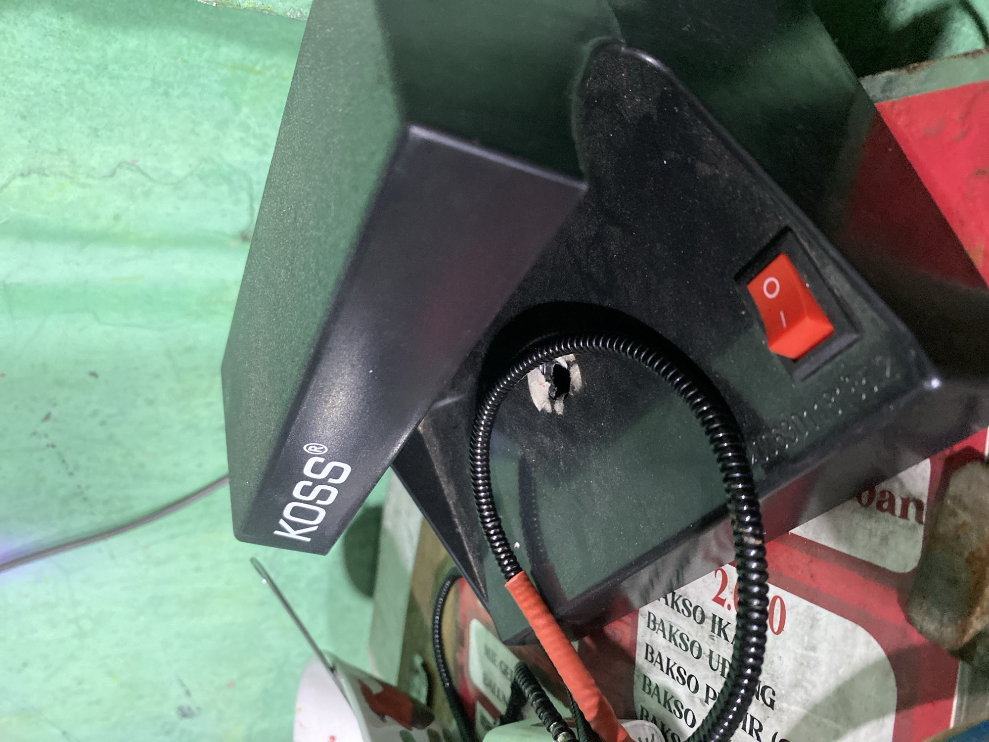 This is a close-up, low-angle, eye-level shot of a black Koss brand electronic device. The device is on a surface that appears to be made of rough, textured green material in the background. In the foreground, there is a black, coiled cable connected to the device, with a section wrapped in red tape. A red rocker switch with a white circle and line symbol is visible on the device. Some text is visible on a white and red patterned surface underneath the device, including "BAKSO". The image is taken in Kasihan, Indonesia.