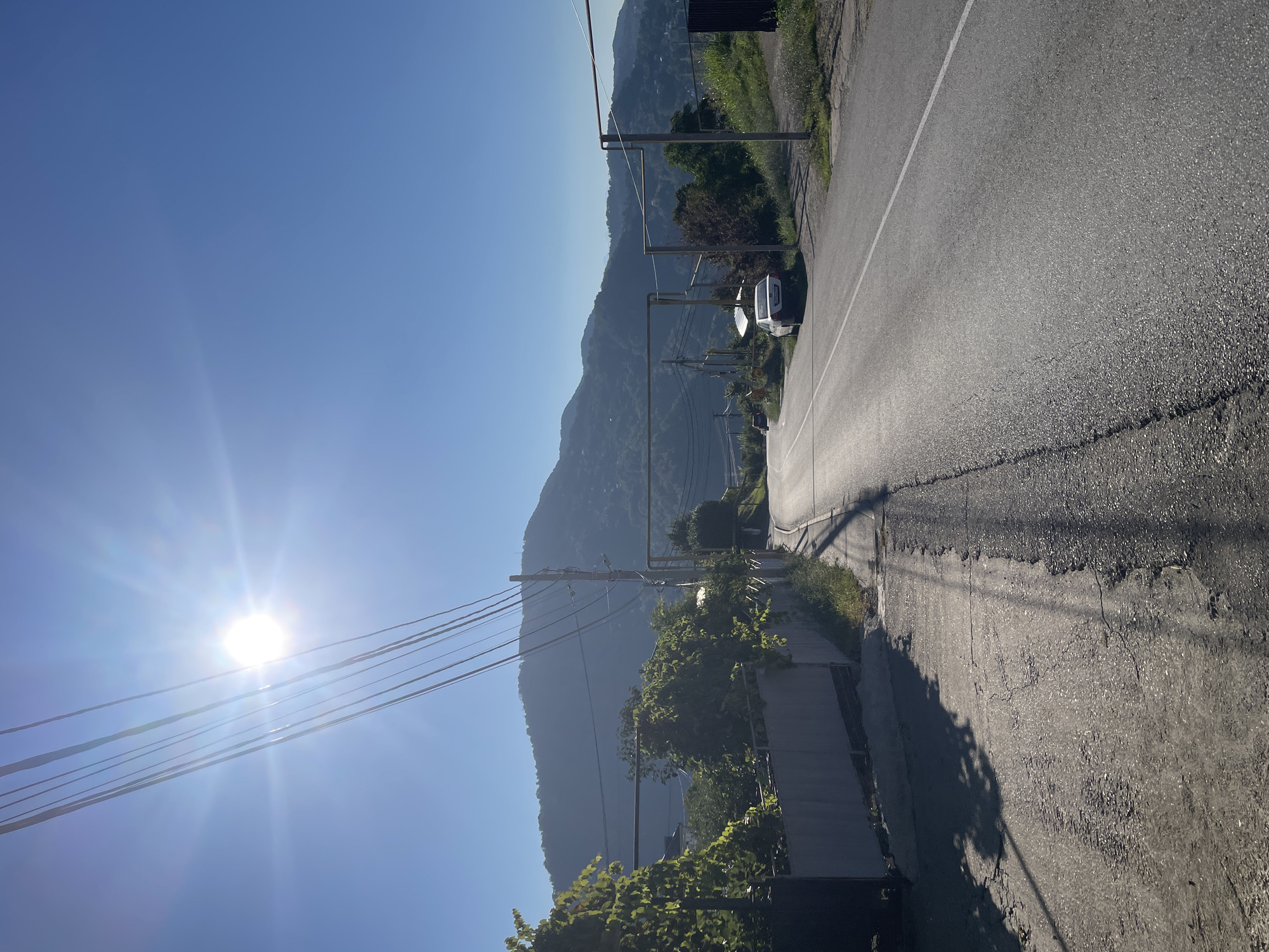 The image depicts a sunlit, paved road curving through a hilly landscape. In the foreground, the road is edged by a gray, low concrete wall on the left, with lush green vegetation growing over it. The road surface shows signs of wear and cracks. Power lines stretch diagonally across the upper left of the frame, with the sun shining brightly through them. In the midground, a white car is parked on the right side of the road, near a small cluster of buildings and trees. Beyond the road, rolling green hills covered in vegetation rise up, forming a mountainous backdrop under a clear blue sky. The overall scene suggests a rural or semi-rural setting in Baranovka, Russia, possibly during daytime.