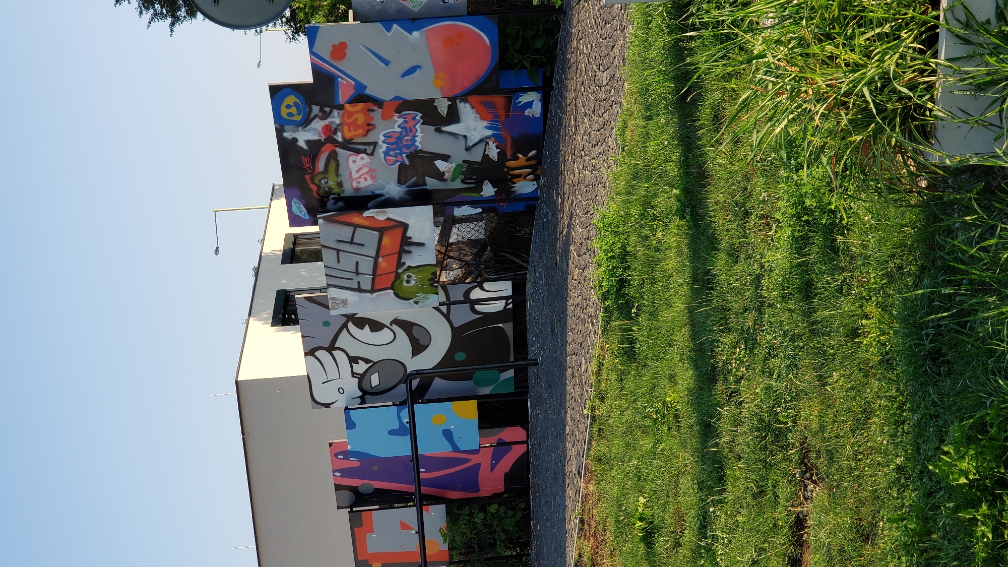 The image depicts a section of a public space with a vibrant graffiti art display. The graffiti is painted on numerous individual panels, creating a mosaic-like effect. The art features cartoonish characters, abstract designs, and text, rendered in a variety of bright and bold colors. Behind the graffiti, a modern, light-colored building with dark-framed windows is visible. The area in front of the graffiti is paved with cobblestones, and a well-maintained lawn with green grass stretches into the foreground. The sky above is clear and blue. The scene suggests an outdoor art installation or a designated graffiti zone within the city.