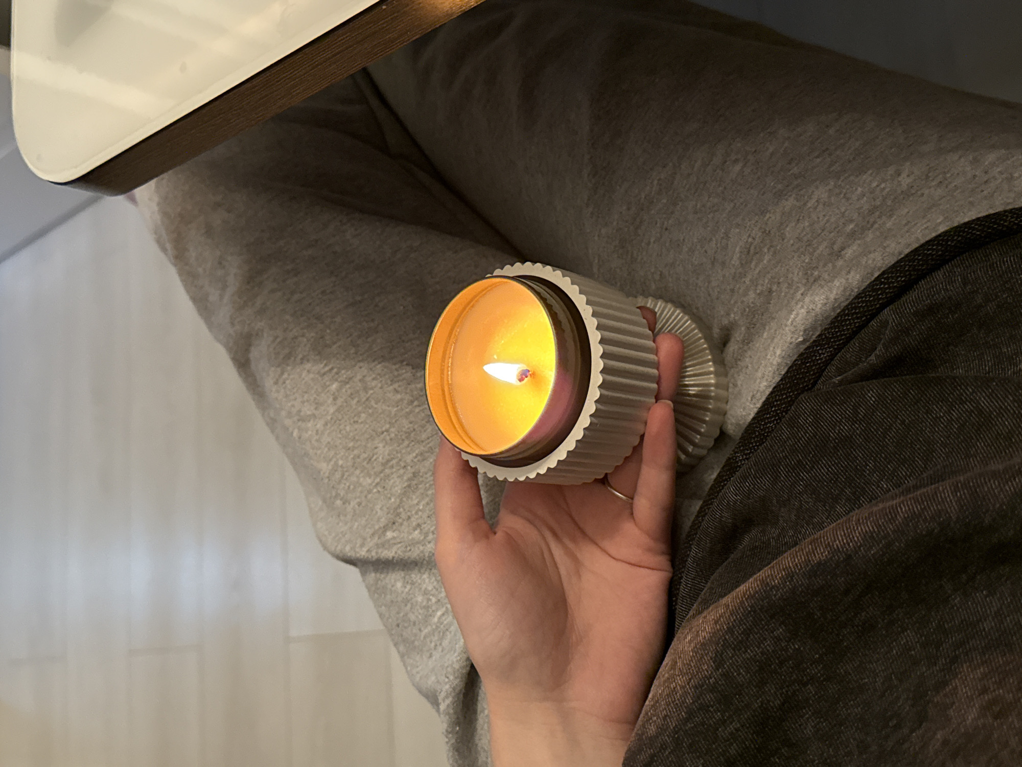 A high-angle, medium shot shows a lit candle in a decorative silver container, held by a person seated on a surface covered in gray clothing. The person's hand is visible, supporting the candle. The clothing beneath the hand is gray sweats and brown corduroy. The flame of the candle is bright and centered within the container. A table with a white top and wooden edge is visible in the upper left of the frame. The background includes a white wall and flooring. The location is unknown.