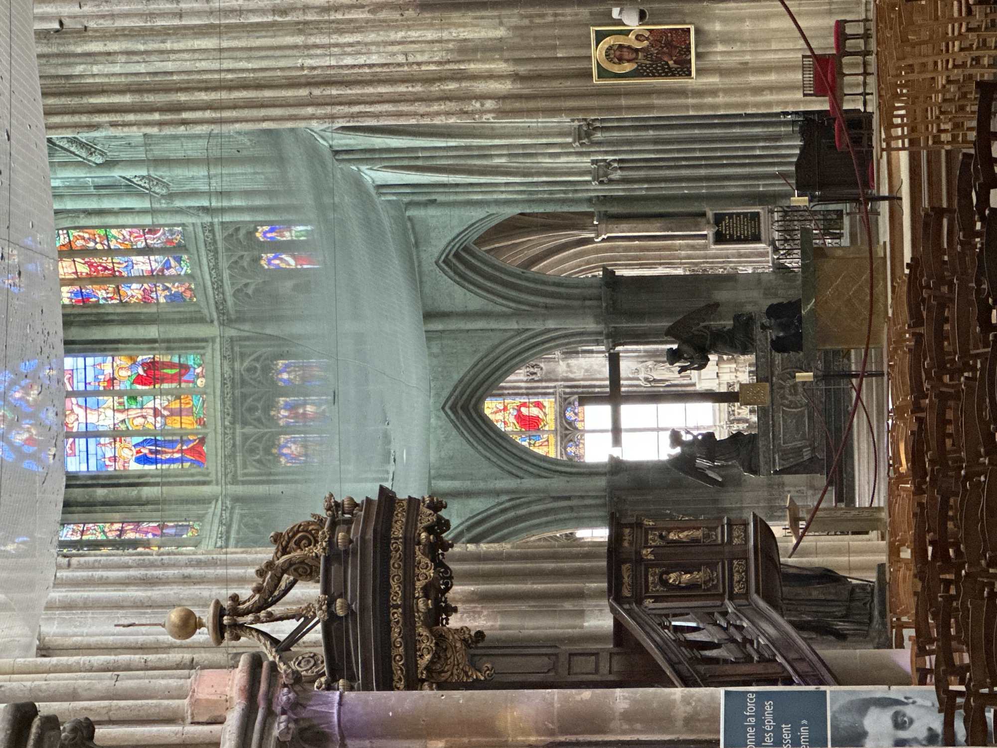 The interior of the Saint-Jacques church in Dieppe, France, is depicted in this image. A large, ornate wooden pulpit stands prominently in the foreground on the left, with intricate carvings and a golden finial. Rows of wooden chairs are arranged on the floor, facing towards the altar area.
The background reveals the grand architecture of the church, characterized by high vaulted ceilings, stone pillars, and arched windows. Several stained-glass windows are visible, featuring colorful religious scenes. Two statues of angels are positioned near the altar, which is partially obscured by a marble-topped structure. A framed icon of the Virgin Mary and child hangs on a pillar to the right. A poster with text and a black and white portrait of a man is attached to a pillar on the far left. The overall scene is illuminated by natural light filtering through the windows and ambient interior lighting.