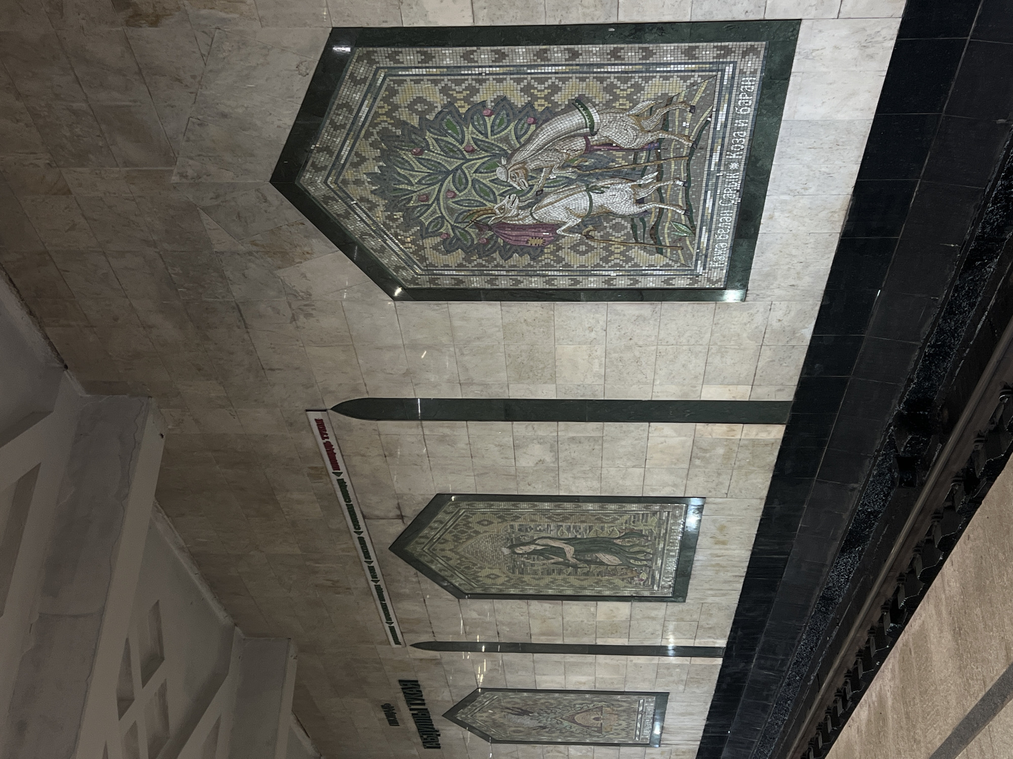 The image shows mosaic panels on the ceiling of the Kazan Kremlin metro station in Kazan, Russia. The panels depict animals, plants, and geometric patterns in a style reminiscent of traditional Tatar art. The ceiling is tiled with light beige marble. There are black rectangular panels separating the mosaics. A safety strip with text in Russian and Tatar is visible on the left. A portion of the wall with black decorative features is seen to the right.