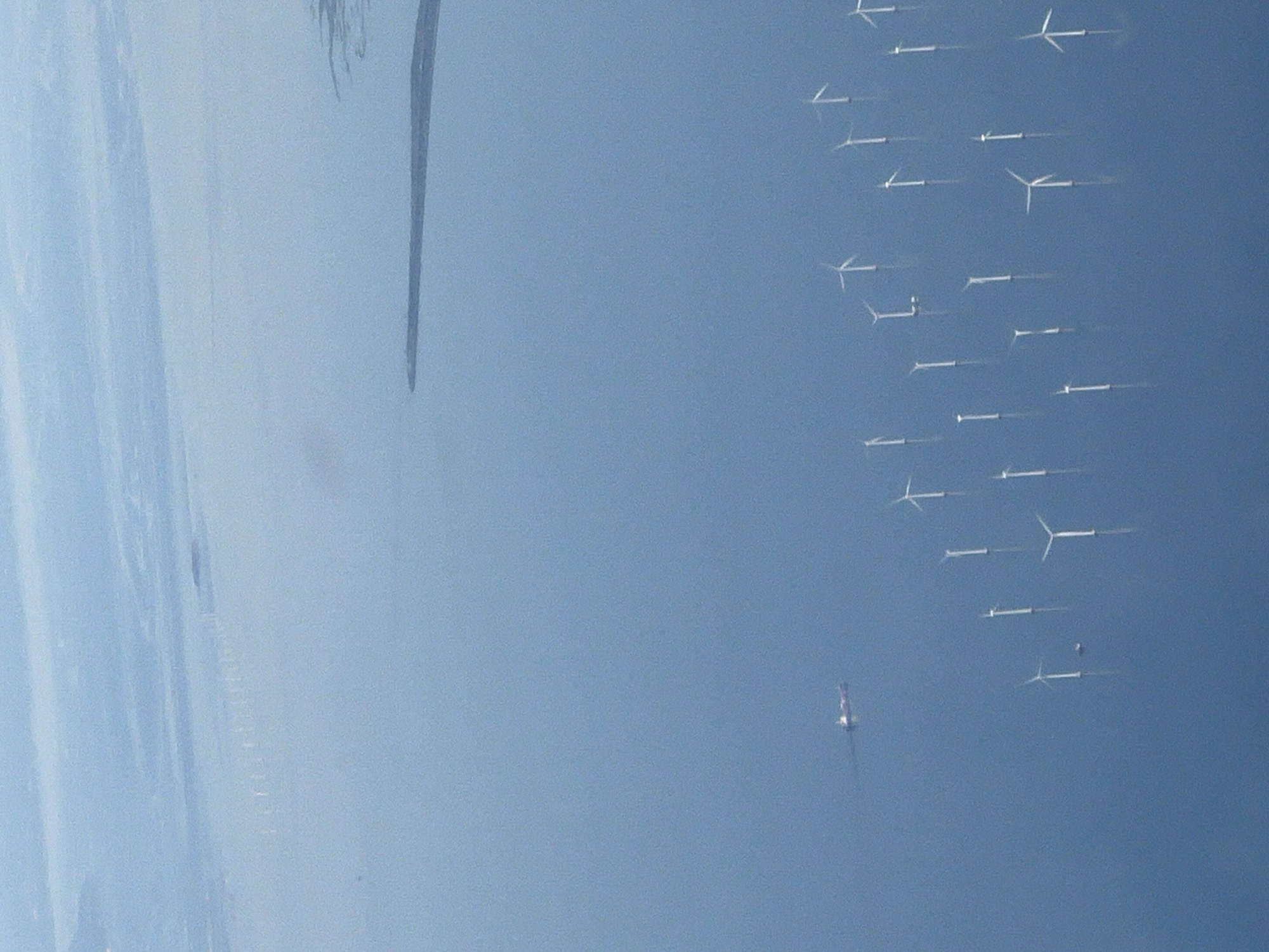 The aerial view depicts a vast expanse of calm, light blue water, characteristic of the Øresund strait off the coast of Kastrup, Denmark. A prominent feature is a large offshore wind farm, with numerous white wind turbines dotting the water's surface. These turbines are spaced out, with some appearing closer to the foreground and others further in the distance, creating a sense of scale and depth. The turbines' blades are varied in their position, suggesting a gentle breeze.
In the mid-ground, a long, dark, landmass or sandbar extends into the water from the right side of the frame. This geological formation contrasts with the uniformity of the water.
Towards the left side of the image, a single cargo ship is visible, a dark silhouette against the blue. It appears to be navigating through the water.
In the upper portion of the image, a hazy, lighter blue area suggests distant land or the sky meeting the water, possibly with a faint suggestion of more wind turbines in the far distance, partially obscured by atmospheric conditions. The overall impression is one of a serene, industrial seascape, highlighting renewable energy infrastructure.