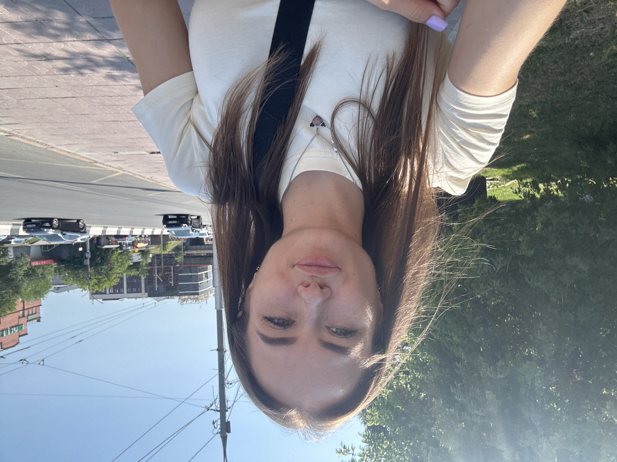 A young woman is taking a selfie outdoors. She is wearing a light-colored t-shirt and has long, brown hair. A thin silver necklace with a heart-shaped pendant is visible on her chest. Her fingernails are painted a light purple color. The background shows a street with cars and buildings, as well as trees and a clear blue sky, suggesting it is daytime and the weather is fair. The image is oriented upside down.