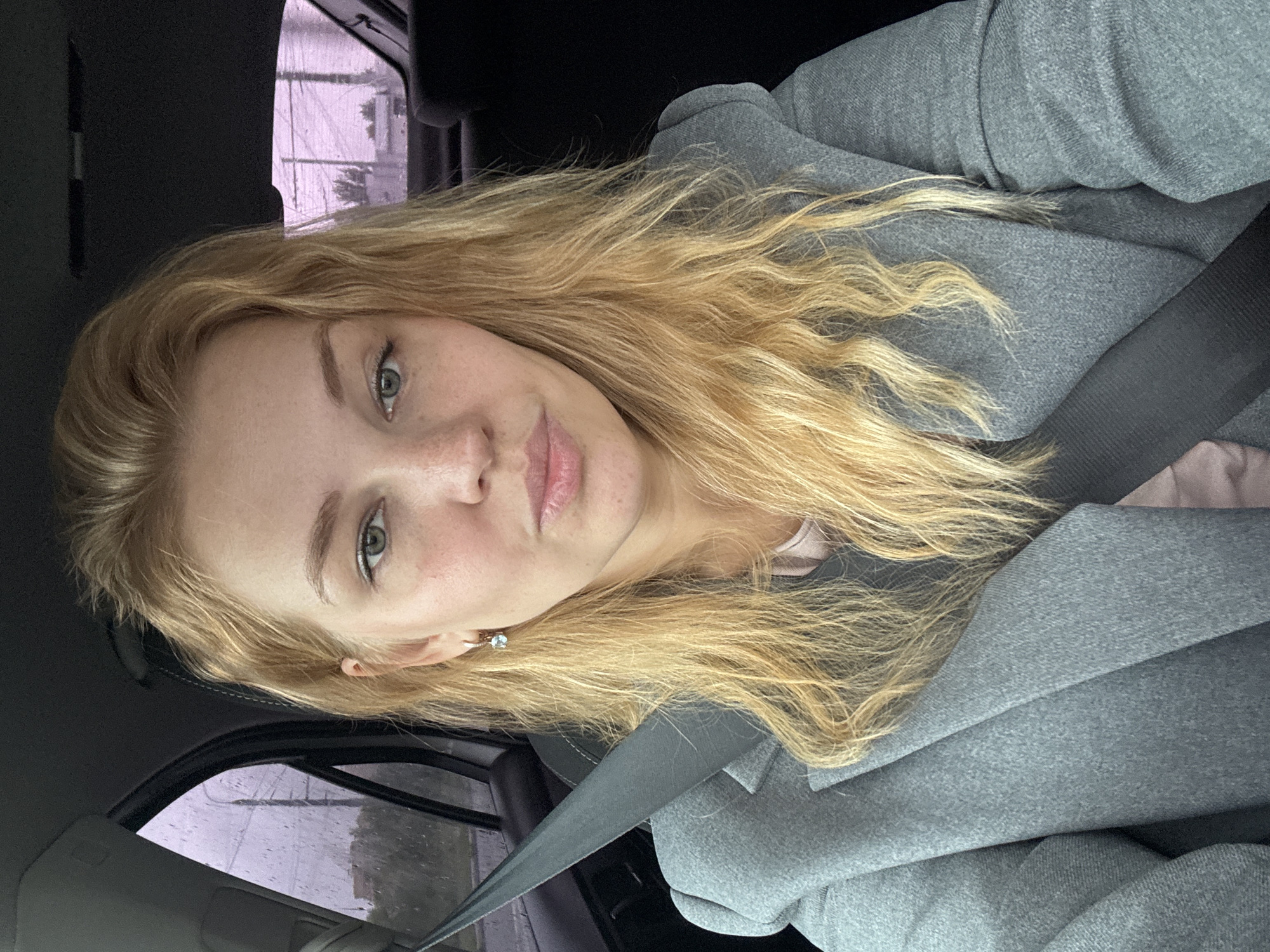 A medium-shot, indoor selfie shows a fair-skinned young woman with blonde, wavy hair, seated in the driver's seat of a car. She is wearing a grey blazer over a pink shirt and a grey seatbelt is diagonally across her body. Her makeup is subtle, with light foundation, mascara, and lip gloss. She is looking directly at the camera with a neutral expression. Rain is visible on the car window behind her, suggesting an overcast or rainy day. The background shows the interior of a car, with the passenger seat and headrest visible. The location is identified as Ignatovo, Russia, though this cannot be visually confirmed from the image itself.