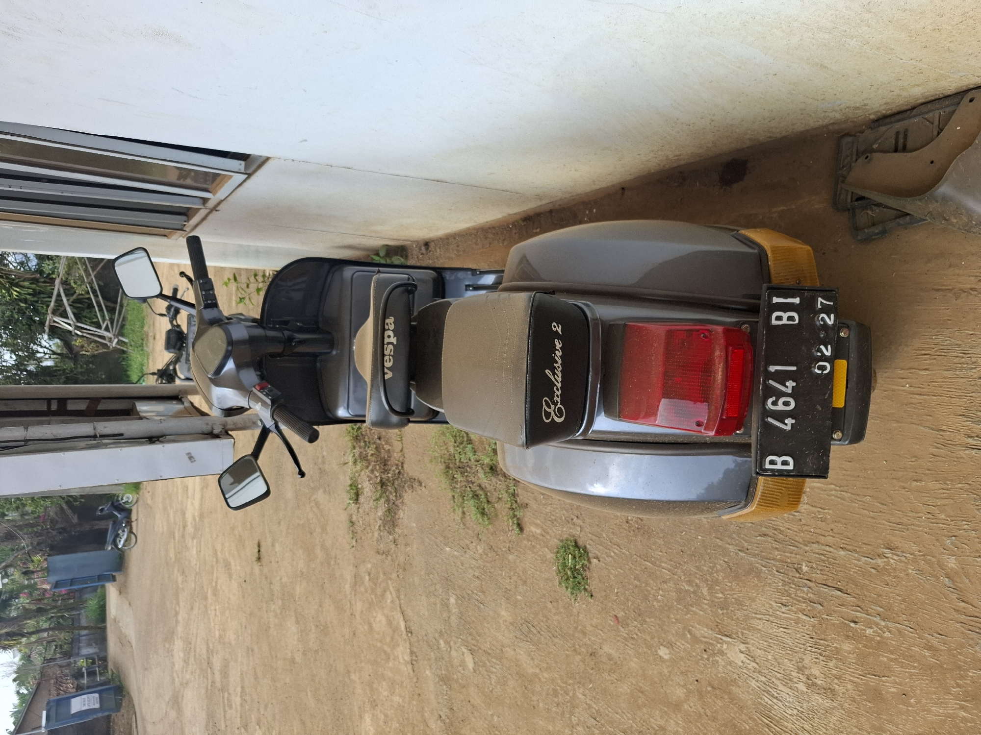 The image shows the rear view of a grey Vespa scooter parked on a dirt and concrete surface next to a white wall. The scooter has a black license plate that reads "B 4641 BI" and "02-27". The seat of the scooter is brown and there is a red tail light and yellow turn signals. The background shows a dirt driveway with some grass and vegetation, and in the distance, a building with blue doors and another scooter. The scene appears to be in a rural or semi-rural area.