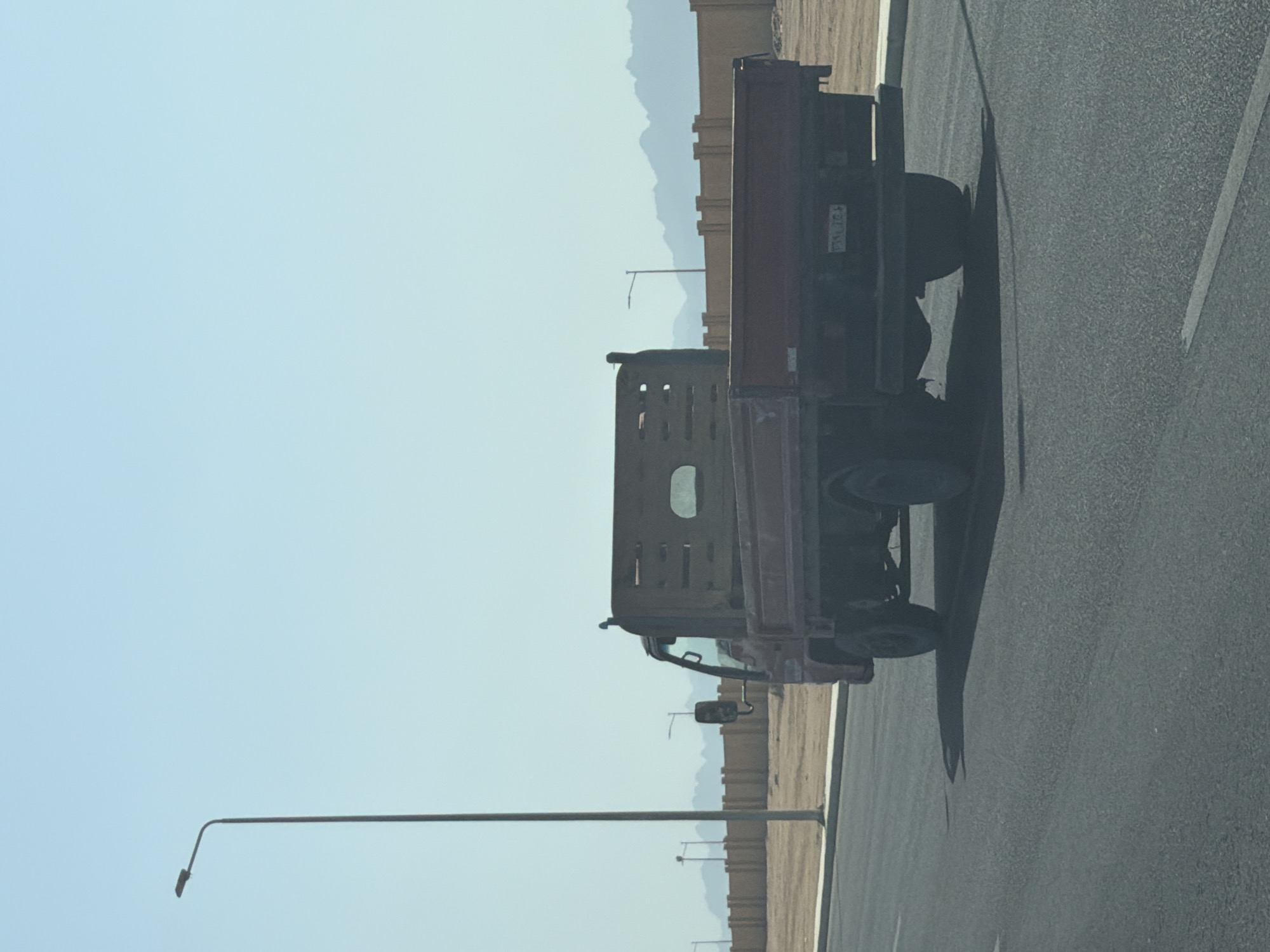 In Sharm El Sheikh, Egypt, a red flatbed truck drives on a paved road under a clear blue sky. The truck is viewed from the rear and slightly to the left. It has a wooden bed with horizontal slats and a small rectangular window in the middle. A license plate is visible on the back of the truck. To the left of the truck, a tall, dark lamp post stands near the road's edge. In the background, a beige concrete fence runs along the edge of an undeveloped, sandy area with some vegetation. Beyond the fence, distant mountains are visible against the horizon. The scene appears to be an outdoor, daytime environment.