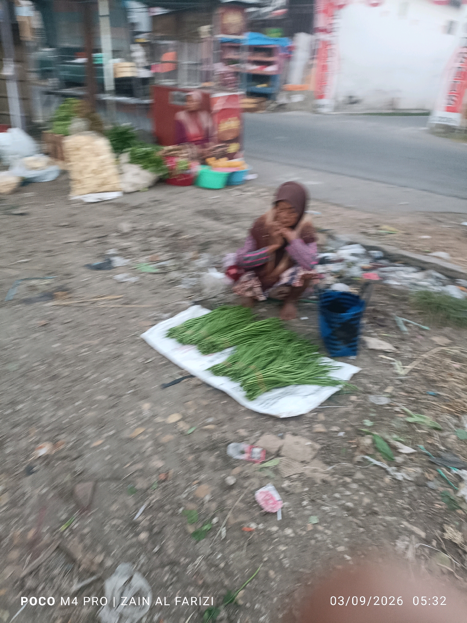 The image depicts an outdoor scene at ground level, likely on a dirt or gravel surface, with a portion of a paved road visible in the background. In the foreground, a young person wearing a headscarf and a patterned garment is crouched down. In front of this individual, a large bundle of green vegetables, possibly leeks or green onions, is displayed on a white sheet or bag. A blue container or bag is placed to the right of the vegetables. The background shows a street with some stalls and structures, suggestive of a market or roadside vendors in Arosbaya, Indonesia. There is some debris scattered on the ground, including plastic bags and wrappers, indicating a less-than-pristine environment. The overall lighting suggests it might be late afternoon or early morning.