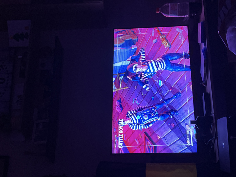 The image depicts an interior scene in the city of Sedlartsi, Bulgaria, characterized by low ambient lighting. A large television screen, positioned centrally, displays a video game identified as "Fortnite: Battle Royale." The screen shows two in-game characters, both rendered in a stylized manner and dressed in black and white striped shirts, black pants, black beanies, and black eye masks, resembling burglars. The male character on the left holds a metallic, box-like object, possibly a safe or device, while the female character on the right strikes a dynamic pose with her arms raised. The virtual environment behind them features a wooden floor with scattered paper currency and other items. A "CONNECTING" message is visible in the lower-left corner of the screen. Below the television, a dark-colored entertainment console or media player is visible, along with a transparent plastic bottle containing a dark liquid on the right. Above the television, a dark shelf is mounted, holding several indistinct objects and a small decorative item resembling a green Christmas tree on the right side. The primary illumination in the room emanates from the television screen itself.