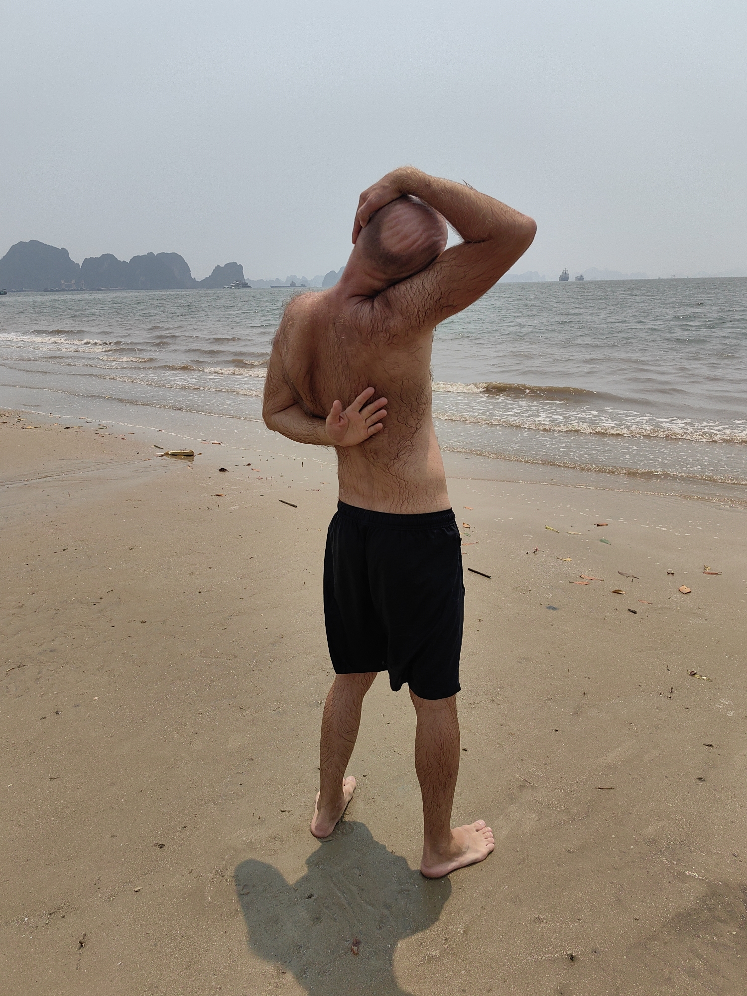 A man stands on a sandy beach facing away from the camera. He has his right hand on his head and his left arm bent backwards with his hand on his back. He is wearing black shorts. The beach stretches towards the ocean, with small waves visible. In the distance, there are islands and boats visible. The location is in Bai Chay Ward, Vietnam. The sky is hazy.