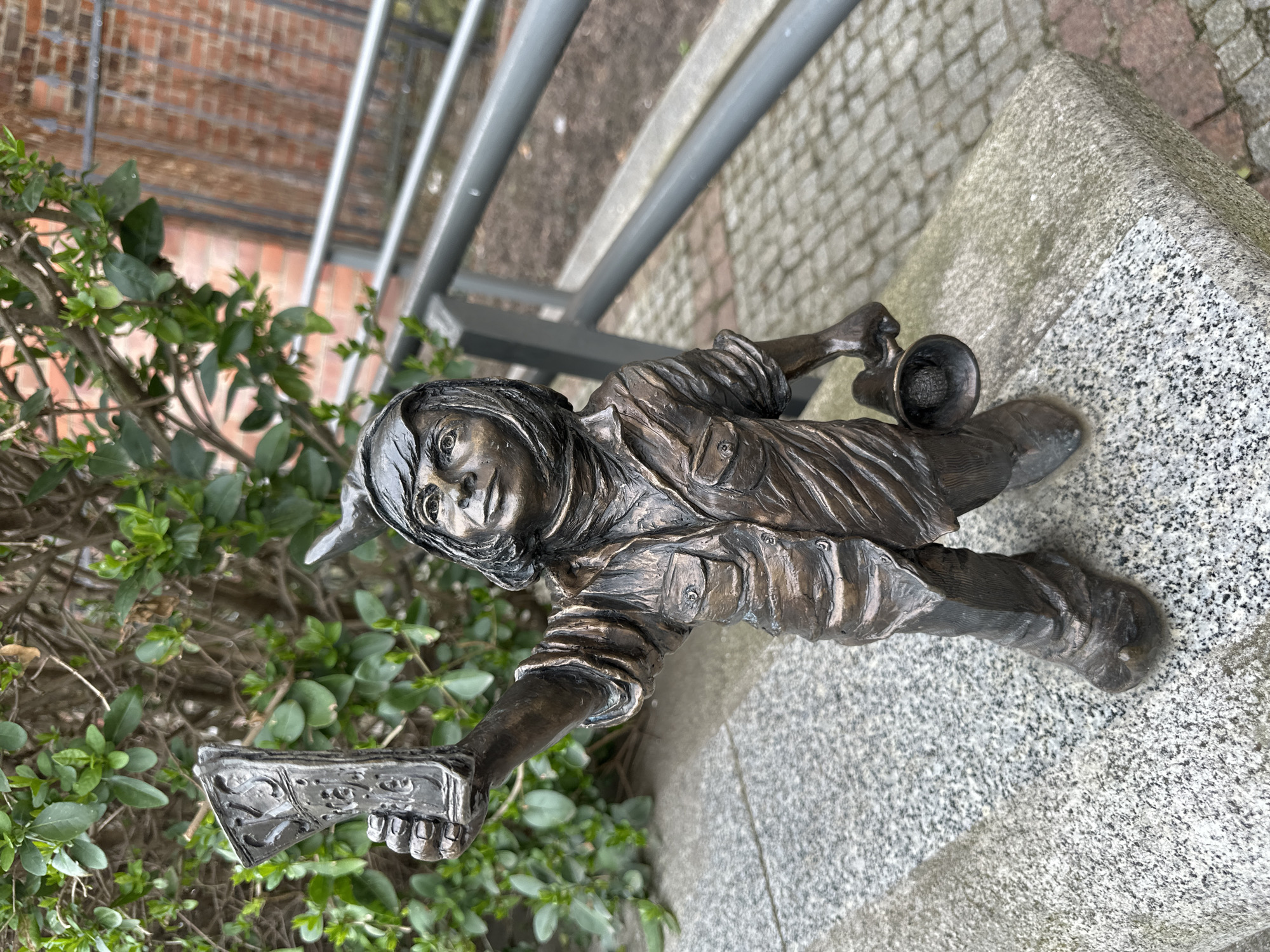 A bronze statue of a dwarf stands near a bush in Wrocław, Poland. The dwarf holds an ax in one hand and a small trumpet in the other. The statue is positioned on a gray stone surface, with a gray metal railing visible behind it. In the background, there is a brick building partially obscured by greenery. The statue is likely one of the many gnome statues scattered throughout the city of Wrocław.