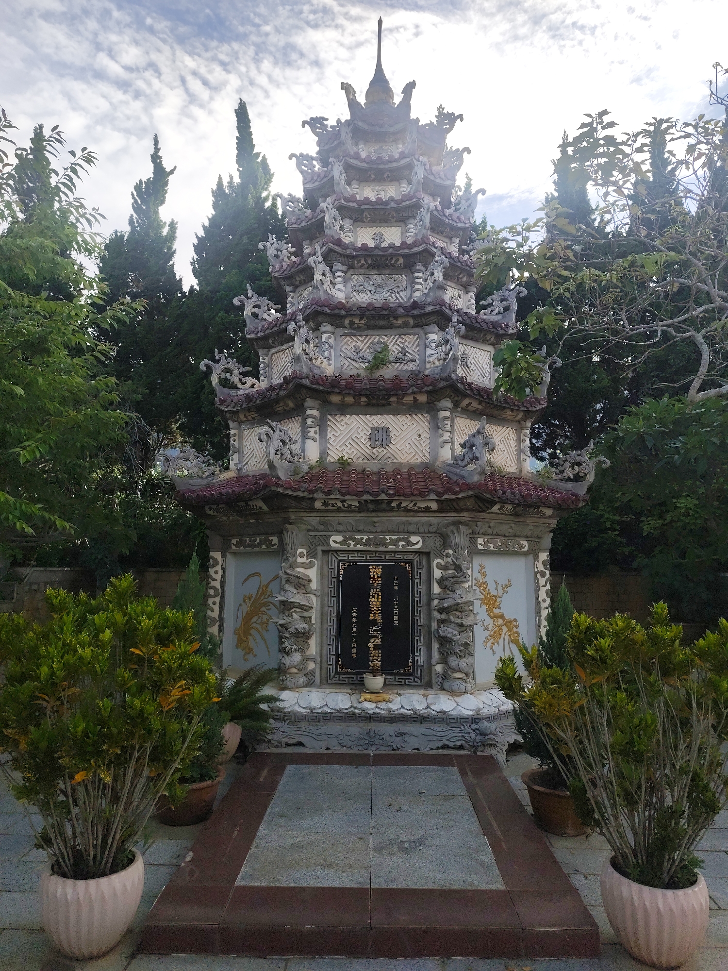 The image shows the tomb of Tran Van Huong, a former Prime Minister of South Vietnam, located in Da Lat, Vietnam. The tomb is a multi-tiered, ornate structure with intricate carvings and decorative roofs. It is surrounded by lush greenery, including trees and potted plants. The sky above is partly cloudy. The tomb is positioned centrally in the frame, with the foreground showing a paved walkway leading up to it. The overall atmosphere appears serene and solemn.