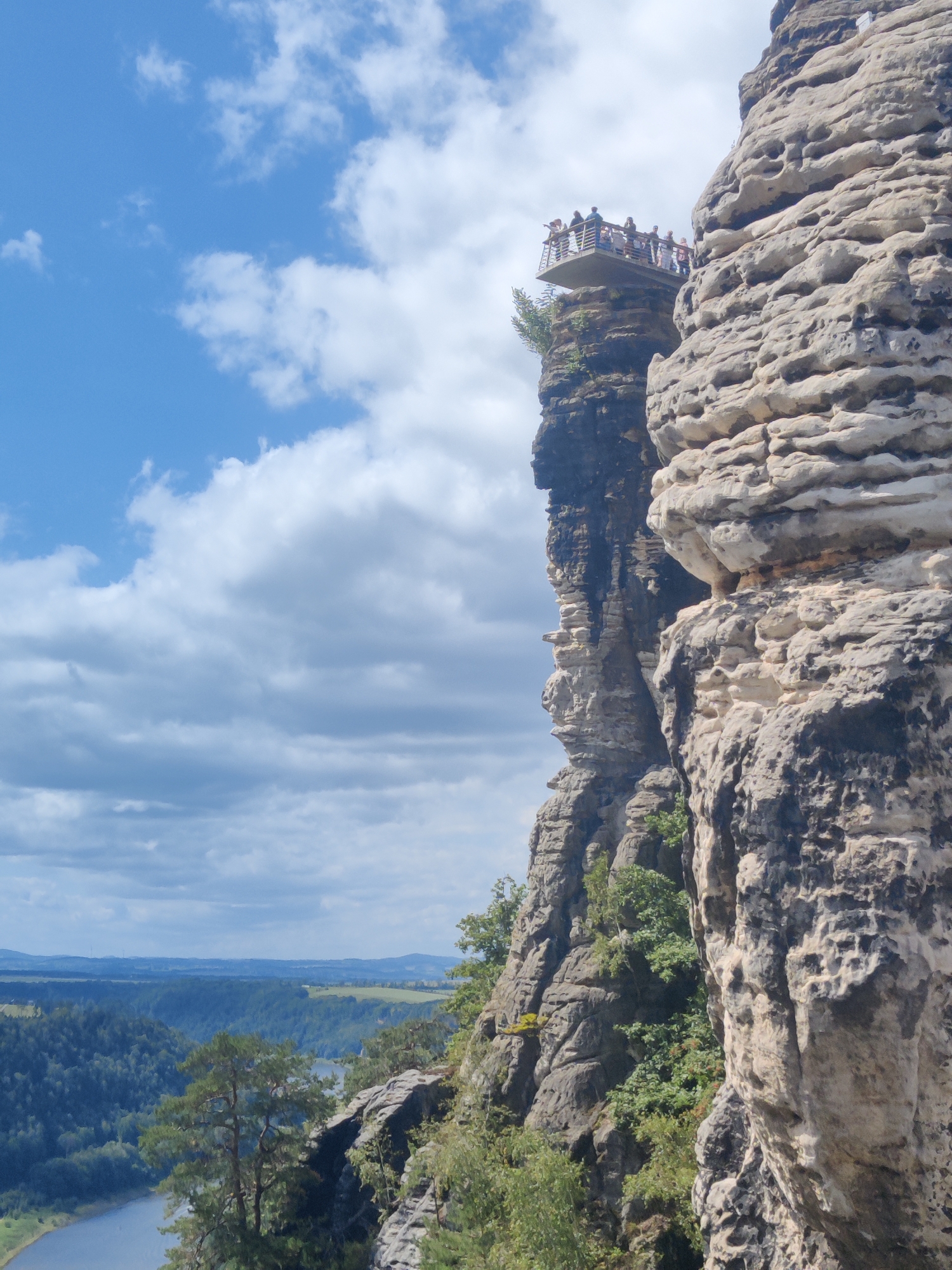 In the foreground, a sheer, weathered rock face dominates the right side of the frame. This rock formation is characterized by its layered texture and numerous natural holes and crevices. To the left, a narrower, darker rock spire rises, with a viewing platform extending outwards from its peak. Several people are visible on this platform, appearing to observe the surrounding landscape. Below the platform, lush green vegetation clings to the rock face. The background reveals a vast, forested valley with a winding river or body of water. The distant landscape is softened by a hazy, blue-green hue, and the sky above is a bright blue with scattered white clouds.