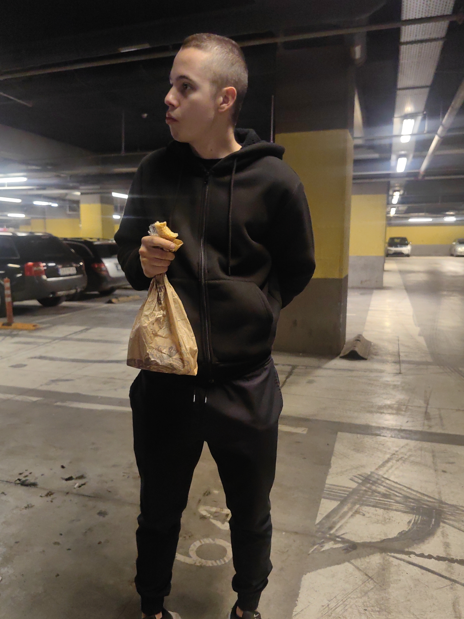 A young, light-skinned male with short hair stands in what appears to be an indoor parking garage. He is dressed in a black hooded sweatshirt and black sweatpants. He is holding a partially eaten piece of food in one hand and a brown paper bag in the same hand. He is looking to his left. The environment is dimly lit with yellow and gray pillars and parked cars in the background. The floor has tire marks and debris. The location could be a parking garage in Sofia, Bulgaria, based on the visual characteristics and architectural style, but there is no definitive information within the image to confirm this.