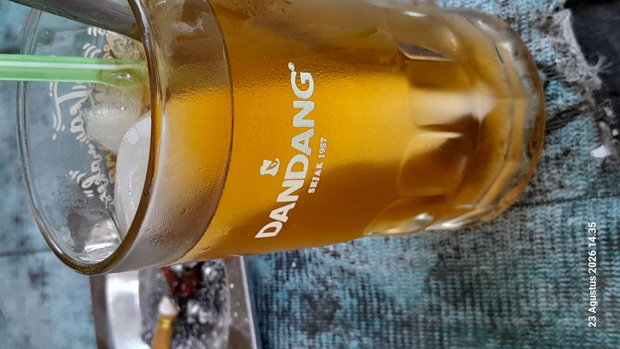 A glass of iced tea sits on a blue, textured surface, likely a tablecloth. The glass, partially filled with amber-colored liquid and ice cubes, has condensation on its exterior. White text on the glass reads "DANDANG SEJAK 1957." A green straw and a metallic spoon are visible inside the glass. To the left, a silver ashtray contains cigarette butts and ash. The background is blurred, showing a blue, textured surface. A timestamp "23 Agustus 2026 14.35" is present in the bottom left corner. The scene appears to be indoors or in a covered outdoor area. There are no people visible in the image. The location specified, Gemaharjo, Indonesia, is not visually verifiable from the image content.