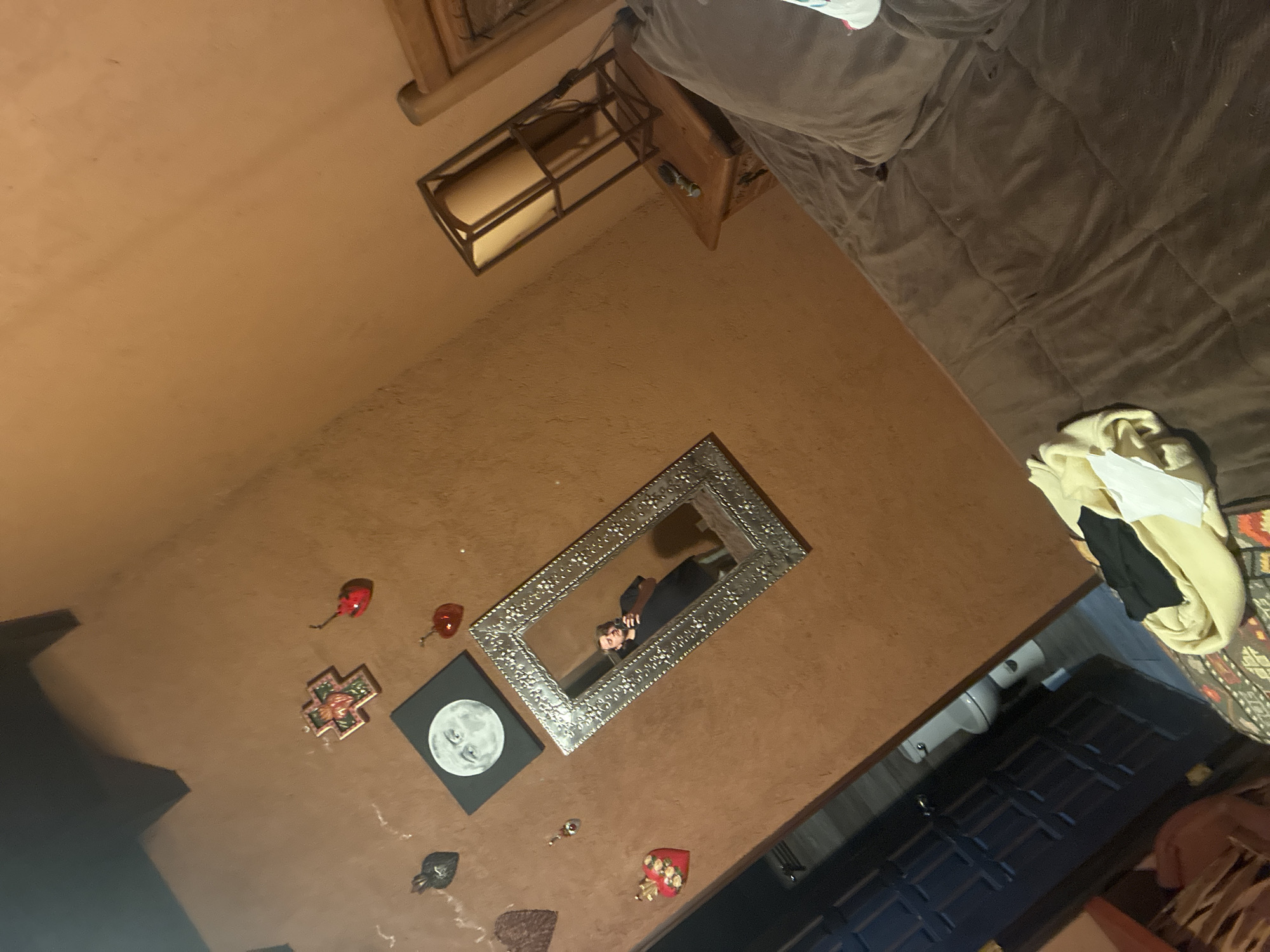 The image shows an interior space. A brown couch dominates the upper right corner. To the left and below, the wall is painted a terracotta color. Decorations on the wall include a rectangular mirror, a painting of a moon, a cross-shaped ornament, and several heart-shaped ornaments. A wooden side table sits near the couch. A black door is partially visible in the lower right corner, next to what appears to be a pile of clothing. The reflection in the mirror reveals a person holding a phone, taking the picture. The location is unspecified but could be Cañada de Madero, Mexico.