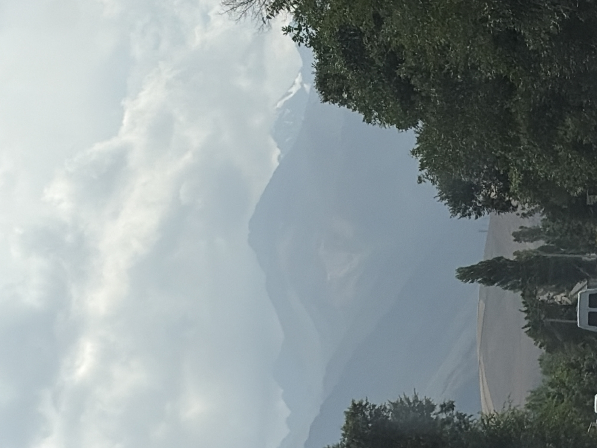 The image depicts a panoramic view of a mountain range under a cloudy sky. The mountains are layered, with the closest ones appearing as dark silhouettes and the farthest ones fading into a hazy blue. Some of the distant peaks show patches of snow. In the foreground, lush green trees dominate the lower portion of the frame, with a hint of a road and a white van visible at the bottom. The overall scene suggests an outdoor setting, possibly a scenic route with mountainous terrain.