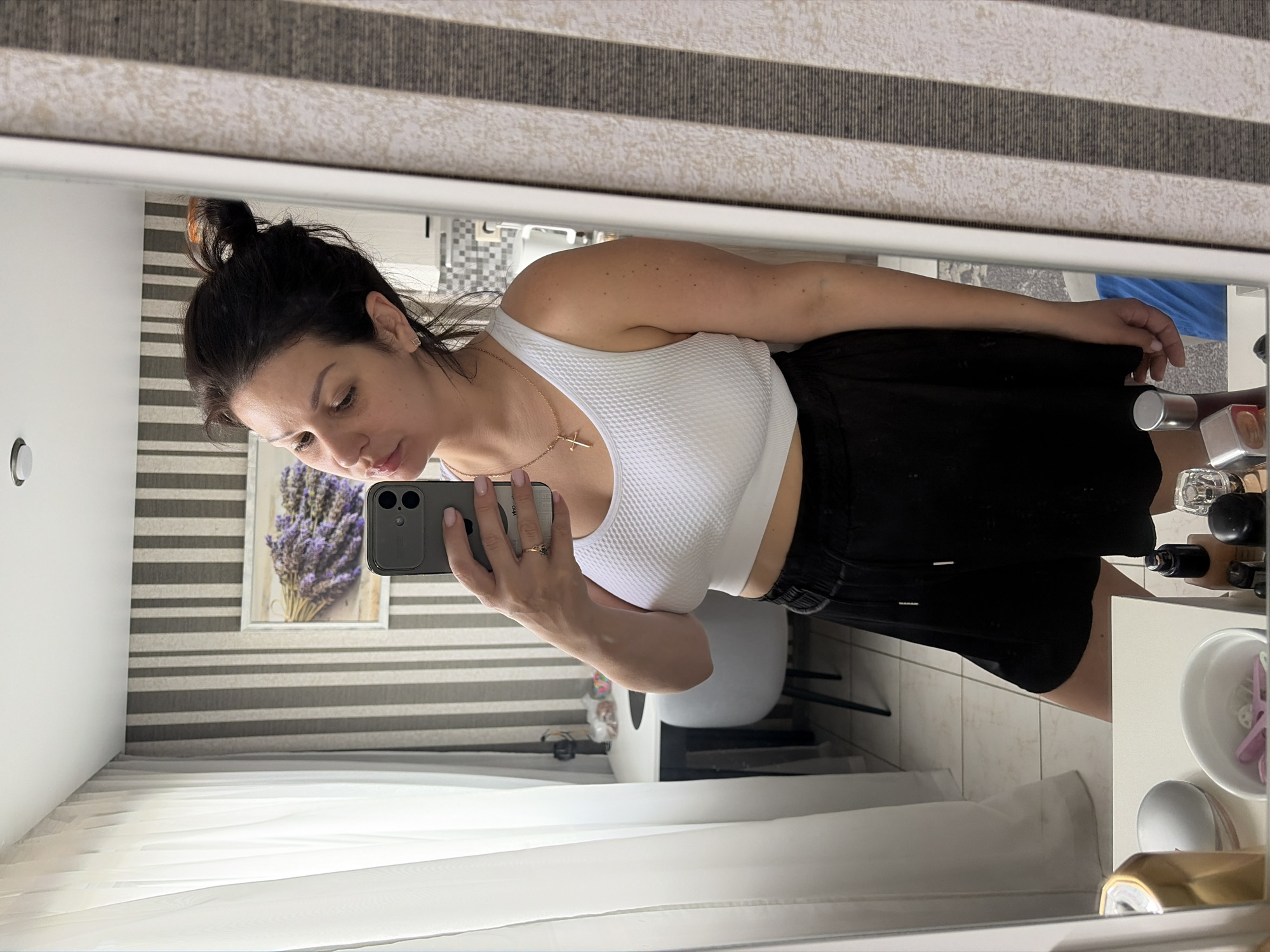 A woman with dark hair pulled up in a bun is taking a selfie in a mirror. She is wearing a white crop top and black shorts. She has on a gold necklace with a cross pendant and a ring on her right hand. The room appears to be a bedroom or dressing area with a white dresser in the foreground holding various items, including a gold can and a white bowl with pink objects. In the background, a desk and chair are visible, along with a framed picture of lavender on the wall. The wall has a striped wallpaper pattern. The lighting suggests it is daytime. The overall scene is indoors. There is no discernible connection to the city of Sochi, Russia.