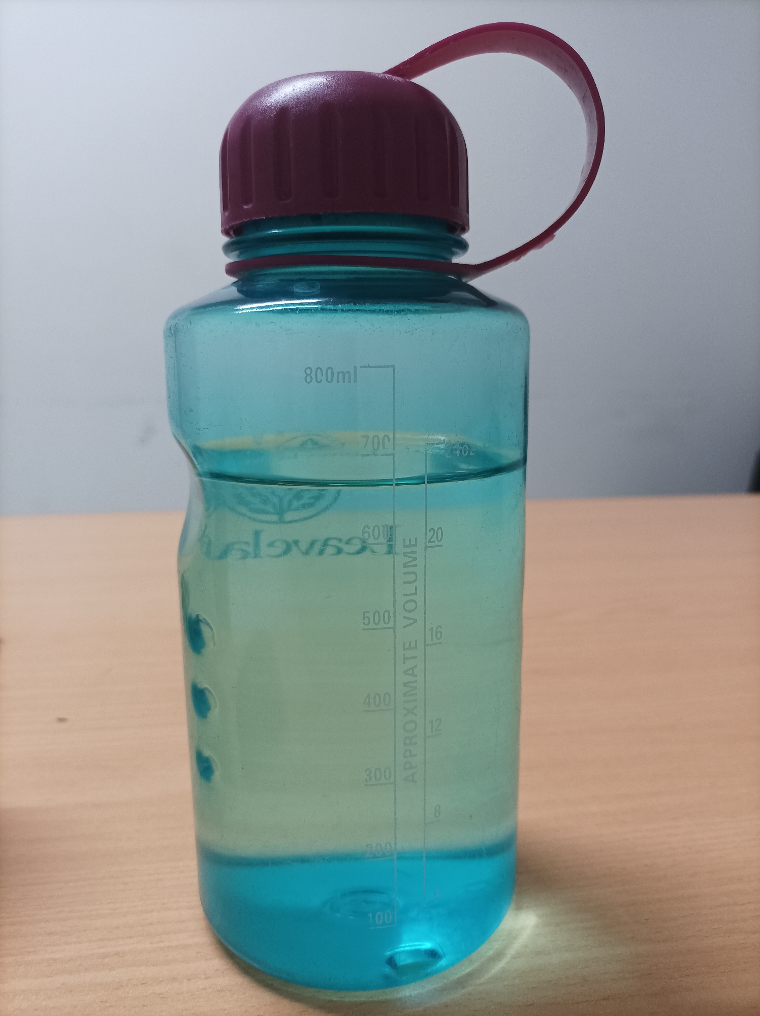 A translucent teal plastic water bottle with a dark red lid and strap sits on a wooden desk. The bottle is approximately two-thirds full of clear liquid, with a blue layer at the bottom. Volume markings are visible on the side of the bottle, including "800ml," "700," "600," "500," "400," "300," "200," and "100," as well as markings in ounces. The background is a plain, light-colored wall. There are no people or animals present in the image. The lighting is even, suggesting an indoor setting. No specific location within Nganjuk, Indonesia, can be determined from the visual information provided.