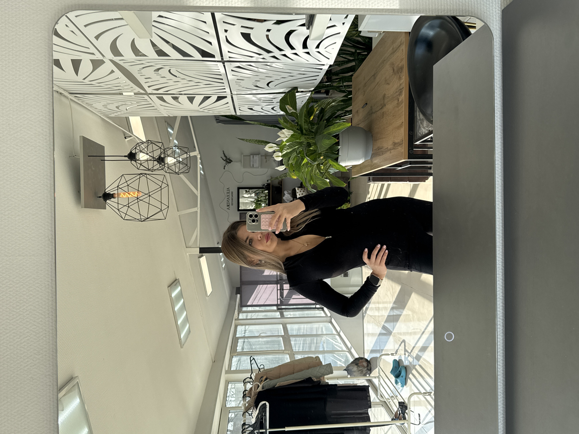 The image, originally rotated 90 degrees counter-clockwise, presents a mirror selfie taken by a woman in a bright, modern interior. A young woman with long, light-colored hair, dressed in a black long-sleeved outfit, holds a smartphone with a pink textured case to capture her reflection. Her right hand rests on her hip, and a smartwatch is visible on her left wrist.
The reflected environment suggests a commercial or public space, possibly a boutique, salon, or office. The ceiling features white, intricately patterned decorative panels. Several modern pendant lights with geometric black frames and exposed filament bulbs illuminate the area. A large potted plant with white flowers, likely a Peace Lily, sits on a wooden surface, next to a dark chair or stool. Text on a wall, mirrored, reads "АЛЕКСАНДРА" and "ЯНТЕХТРАЙТ".
Large windows allow ample natural light, indicating daytime with clear sunshine, as evidenced by shadows cast on the light-colored floor. A white metal clothing rack holding various garments, such as jackets, is visible, along with two bright blue clogs on the floor. The mirror itself is rectangular with rounded corners and features a dark grey frame on the right, which includes a circular touch control button. The wall adjacent to the mirror has a subtle textured wallpaper.