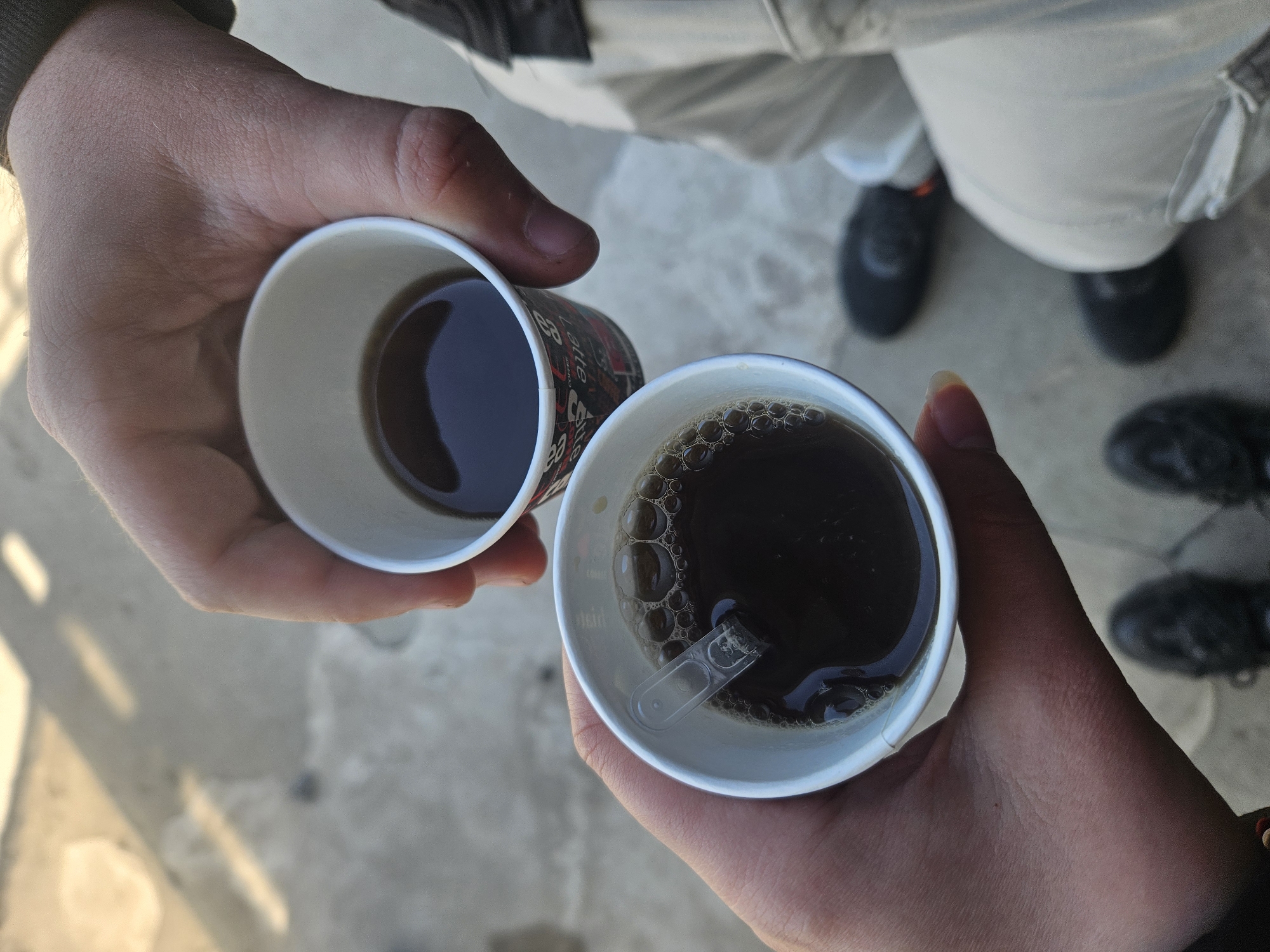 Two hands are holding paper cups filled with a dark liquid, likely coffee or tea, above a concrete surface. In the background, part of a person's body is visible, wearing light-colored pants and dark shoes. The right cup has bubbles on the surface and a clear plastic stirrer inside. The location is unknown, and there is no clear indication that the image was taken in Burgas, Bulgaria.