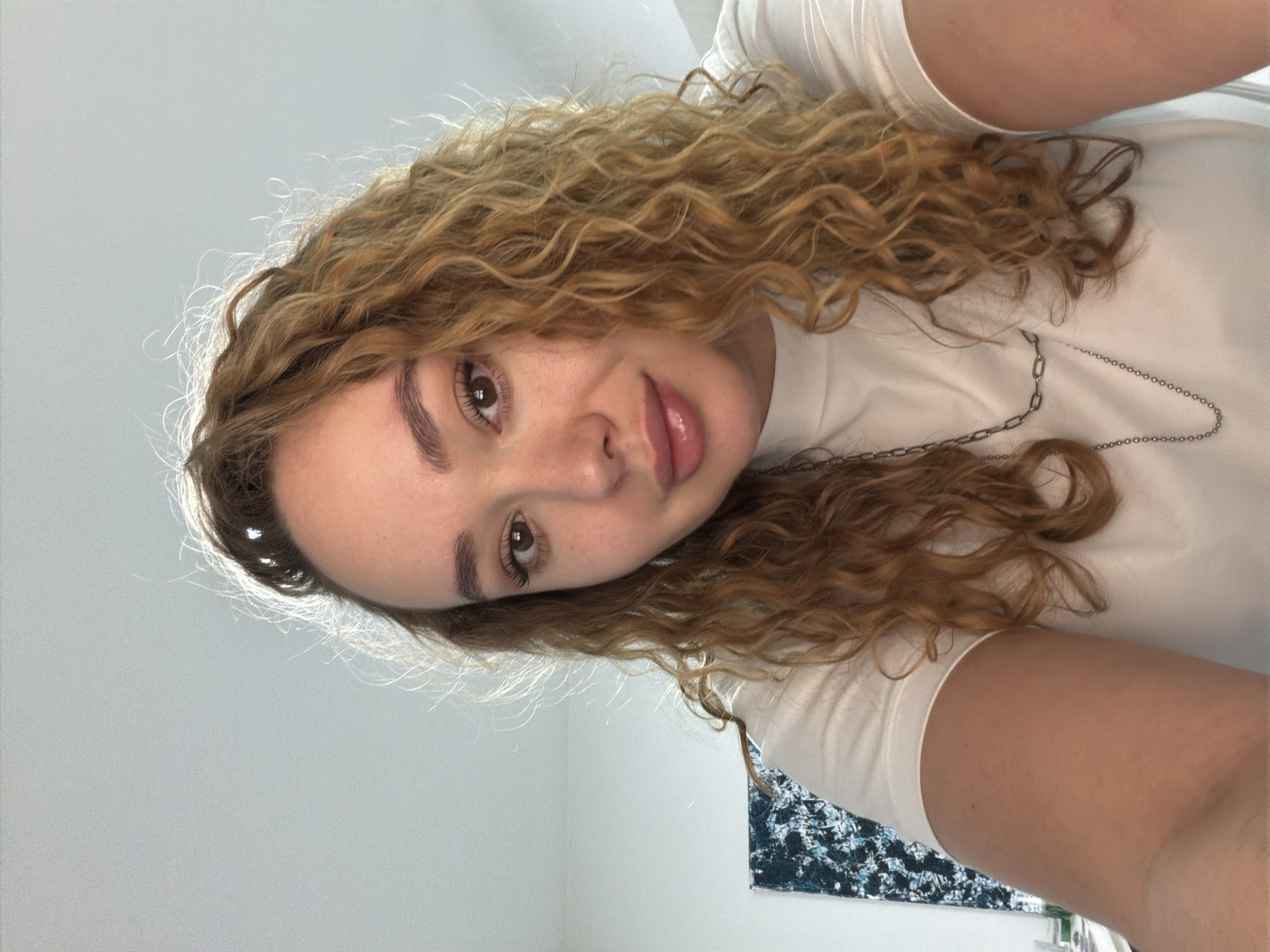 A young woman with curly, light brown hair is positioned close to the camera. She has fair skin, dark eyebrows, brown eyes, and pink lips. She wears a white t-shirt and a silver necklace. The background is a white wall with a dark blue and white patterned object. The photo appears to be taken indoors. There is no evidence to indicate the location of the photo is in Ramat Gan, Israel.