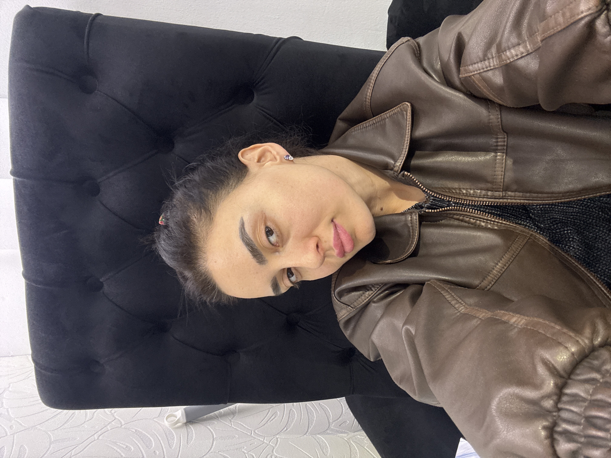 A woman with dark hair and light skin is seated on a black, button-tufted chair. She is wearing a brown leather jacket over a dark top. The background includes a white wall and a patterned white surface. The image was likely taken indoors. The location is unknown.