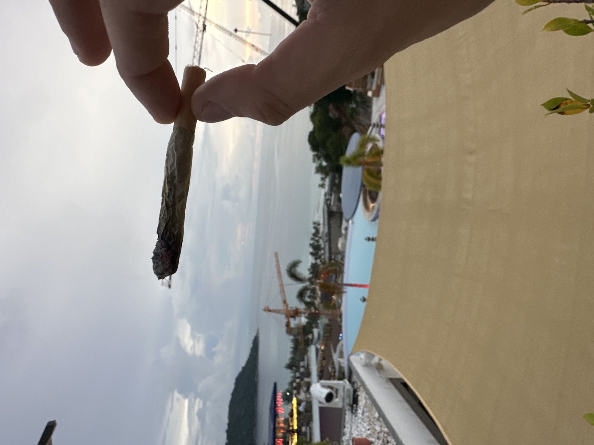 A hand holds a lit marijuana joint, its ash glowing. The joint is held up against a hazy, overcast sky. In the background, a resort setting is visible, with a swimming pool, lounge chairs, and some greenery. Construction cranes are also present, suggesting ongoing development in the area. The lighting suggests it is either early morning or late evening. The scene appears to be in Karon, Thailand, given the location context provided. There is no visible text in the image.