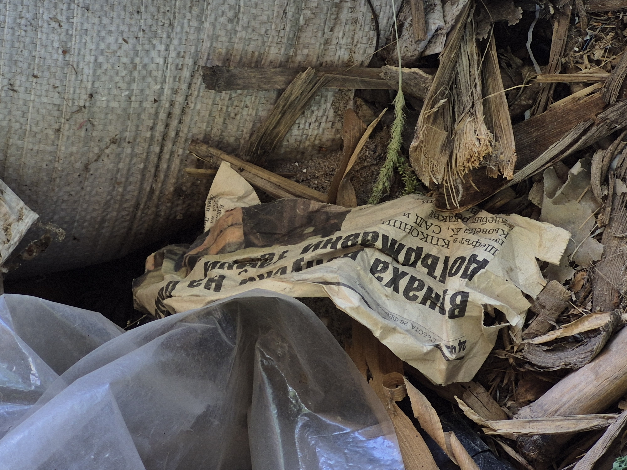 The image shows a pile of debris, including a crumpled newspaper with Cyrillic text, pieces of wood, and a transparent plastic sheet. The newspaper appears to be old and weathered. The debris is situated in an outdoor environment, possibly a back alley or a construction site. The location is identified as Sadina, Bulgaria. There are no people visible in the image. The scene is still and appears to be a snapshot of an abandoned or neglected area.
