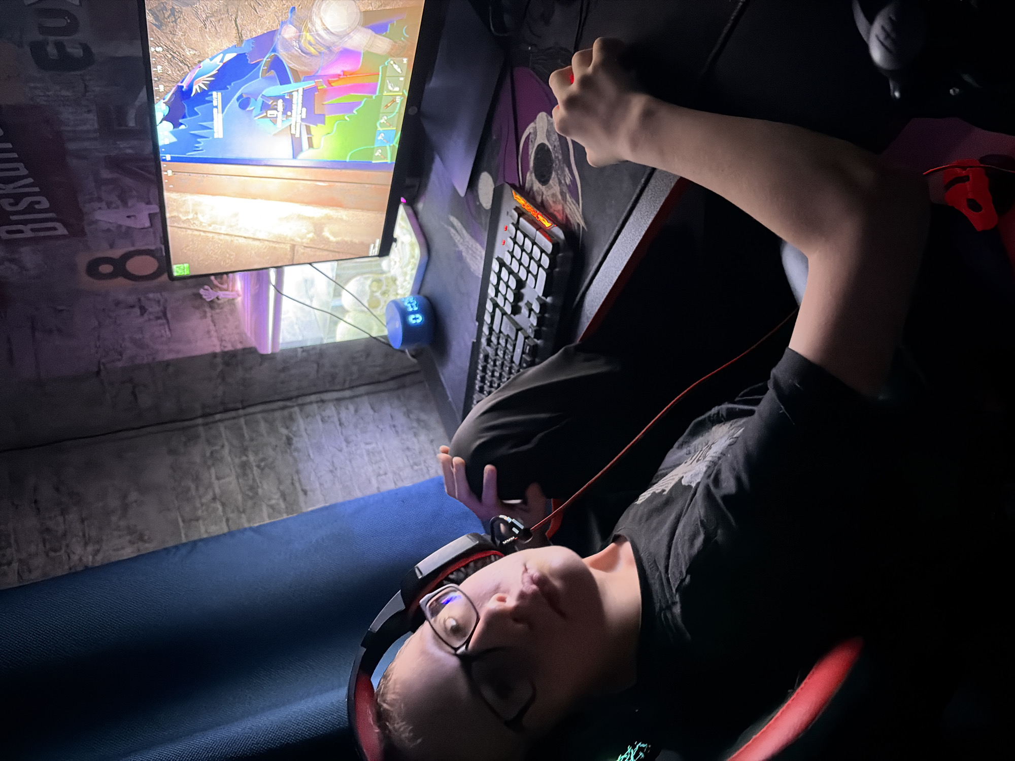An overhead shot captures a young person deeply engrossed in a video game within a dimly lit room. The individual, wearing glasses and black-and-red headphones, is lying back in a gaming chair with red accents, looking intently at a computer monitor that displays a colorful, blocky game environment. Their right hand is positioned on a gaming mouse, which has a red light, resting on a mouse pad featuring a stylized graphic. A black keyboard with some illuminated orange keys is also on the desk.
Behind the person's head is a blue cushion or pillow. On the desk, near the monitor, sits a small blue circular device displaying "08," possibly a clock or timer. The wall to the left of the monitor has a rough, textured finish with visible text, including the word "BISKUIT," the number "8," and partially obscured letters that resemble "EU." The overall ambiance is that of a dedicated gaming setup, likely in the evening or night, enhancing the focus on the screen. The person's expression is one of concentration as they engage with the game.