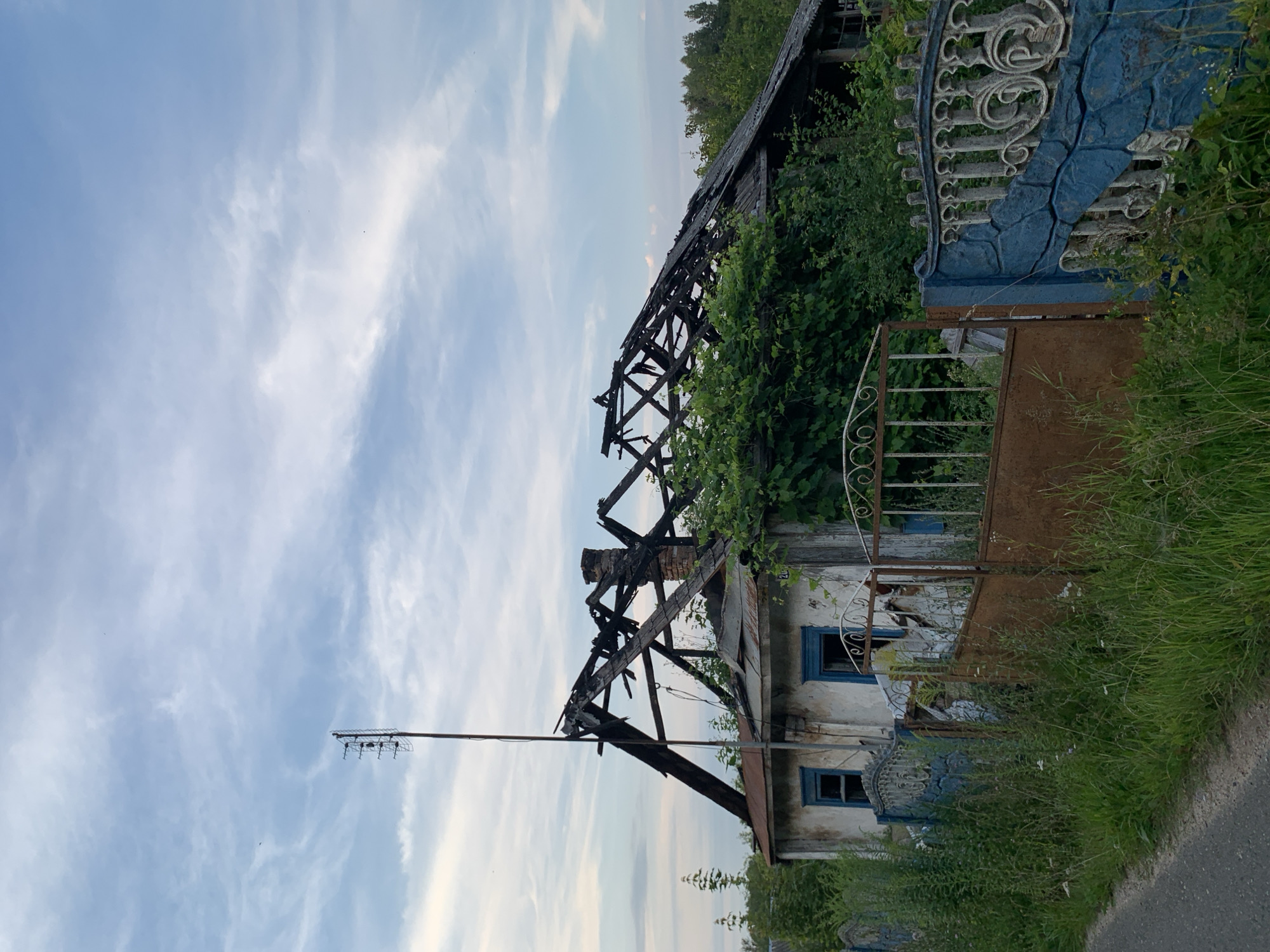 The image shows a severely damaged, abandoned house with a collapsed roof, located in the city of Kapyĺ, Belarus. The structure appears to be a single-story dwelling with white, weathered walls and blue-framed windows. The roof's wooden beams are exposed and charred, indicating a fire. Overgrown vegetation, including vines and tall grass, engulfs the property, reaching up the walls and surrounding the fence. A metal antenna stands tall next to the house. In front of the house is a dilapidated fence with sections of rusty metal and decorative blue stonework. A rusty, open gate stands between two sections of the fence, leading into the overgrown yard. The scene is set against a cloudy, pale blue sky. The overall impression is one of neglect and ruin in a rural or semi-rural setting.