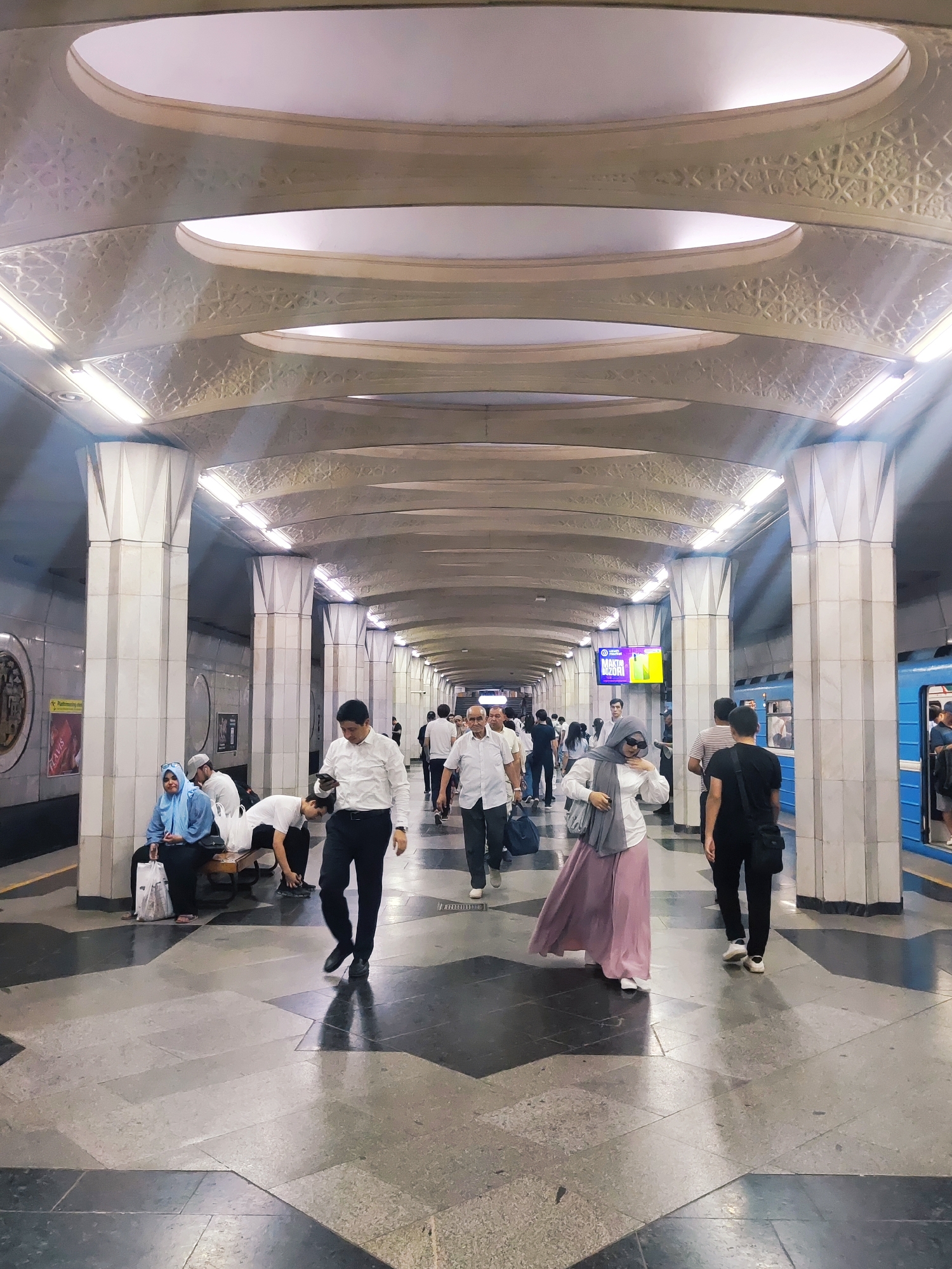 This image depicts the interior of a Tashkent metro station, characterized by its ornate architecture. The station features a long, vaulted ceiling with intricate geometric patterns and recessed lighting, supported by robust, fluted pillars. The floor is a mosaic of dark and light grey tiles arranged in a geometric pattern.
Several individuals are visible, some walking and others seated. In the foreground, a man in a white shirt and dark pants is walking towards the right of the frame, looking at his phone. To his left, a woman in a grey hijab, sunglasses, and a long pink skirt is walking towards the right. Further back, on the left side of the station, a woman in a blue hijab is seated on a bench with bags beside her. Several other people, dressed in casual and formal attire, are dispersed throughout the station, some appearing to be waiting for a train. A blue metro train is visible on the tracks to the right, with passengers boarding or disembarking. The overall atmosphere suggests a typical day in a busy public transportation hub.