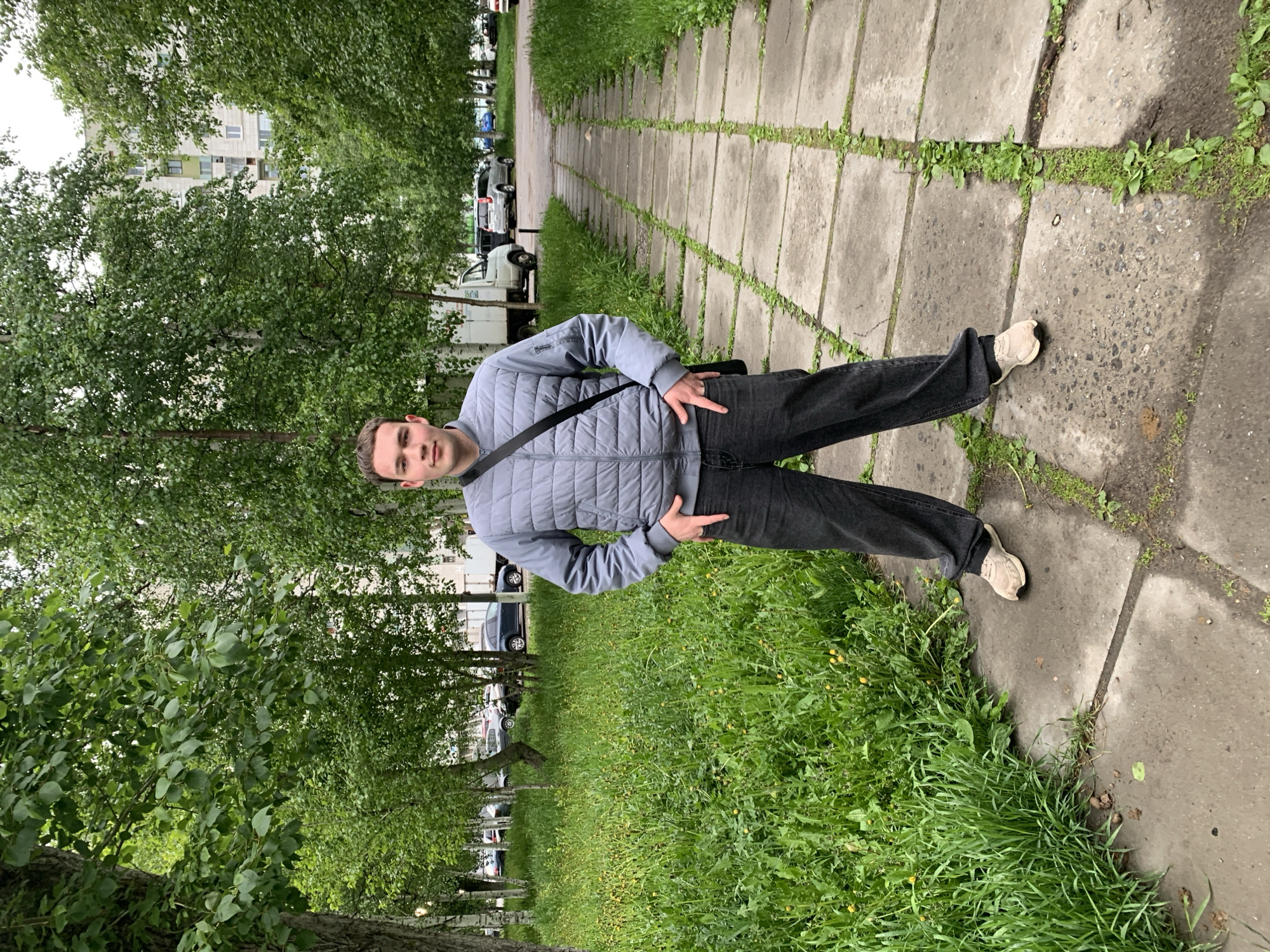 A young man stands centered on a concrete slab path, posing with both hands on his hips, each making a "rock on" hand gesture. He looks directly at the camera with a neutral to slightly smiling expression. He is wearing a light grey quilted jacket with a black cross-body bag, black jeans, and light-colored sneakers. The text "SWED" is visible on the left chest of his jacket.
The path is composed of rectangular concrete slabs, with grass and weeds growing abundantly in the cracks and alongside it. To the left of the path, there's a row of trees, likely birches, lining a grassy area where several cars are parked. Further in the background, multi-story residential buildings are visible under an overcast sky. To the right of the path, there's a wide expanse of overgrown green grass with some yellow flowers. The concrete path and surrounding ground appear damp, suggesting recent rain.
The scene takes place outdoors in what appears to be a residential area or park in Severodvinsk, Russia, during the daytime with cloudy weather.