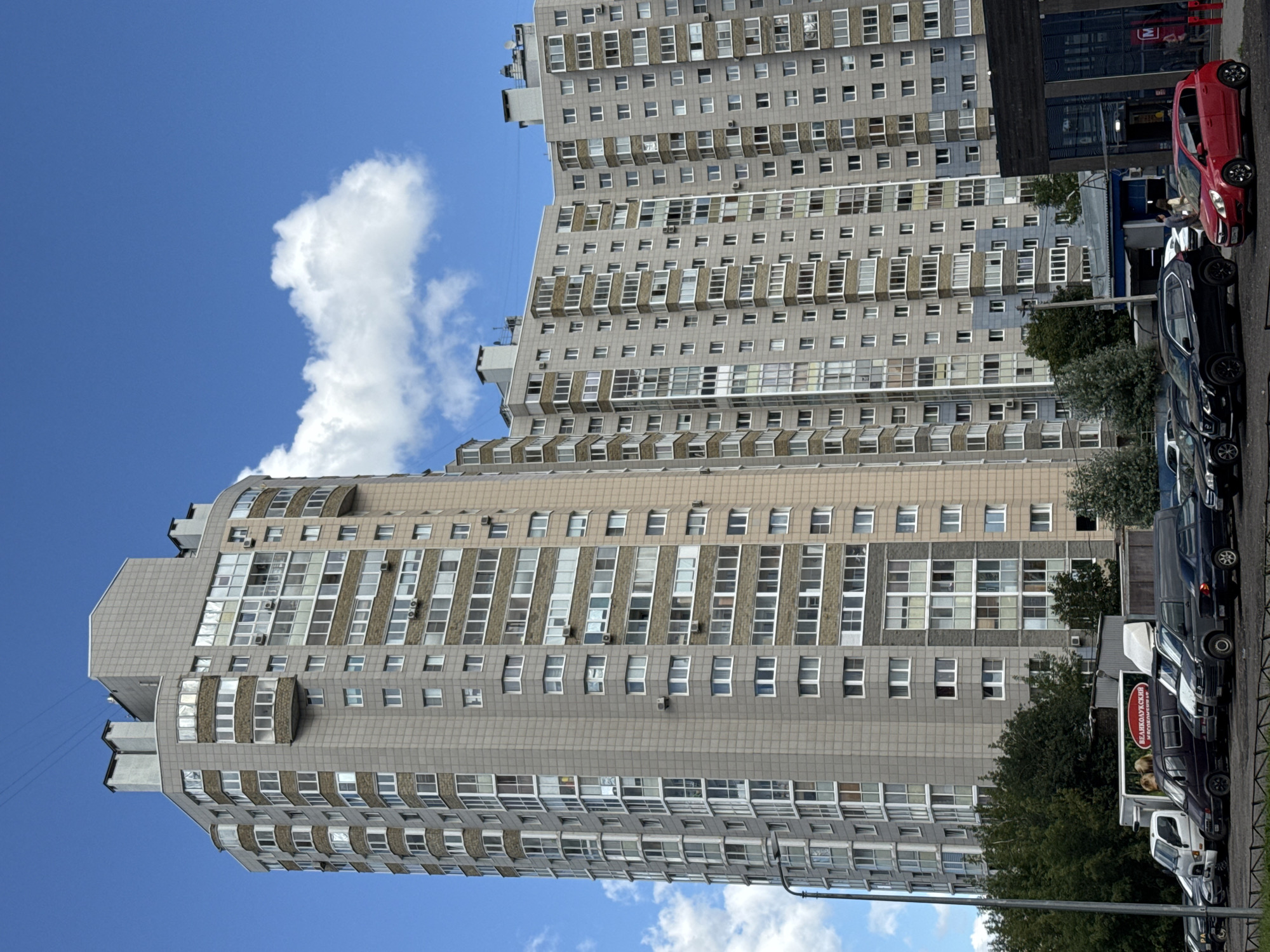 The image shows a modern, tall apartment building with a curved facade, located in Vsevolozhsk, Russia. The building is predominantly light gray with stone-like cladding and features numerous windows, some of which are enclosed balconies. Several smaller buildings and parked cars are visible at the base of the apartment building. The sky is blue with scattered white clouds. A street lamp is present on the left side of the image. In the foreground, a line of parked cars, including vans, SUVs, and a red hatchback, are visible. A building with glass windows and a sign that reads "M" is to the right of the apartment complex.