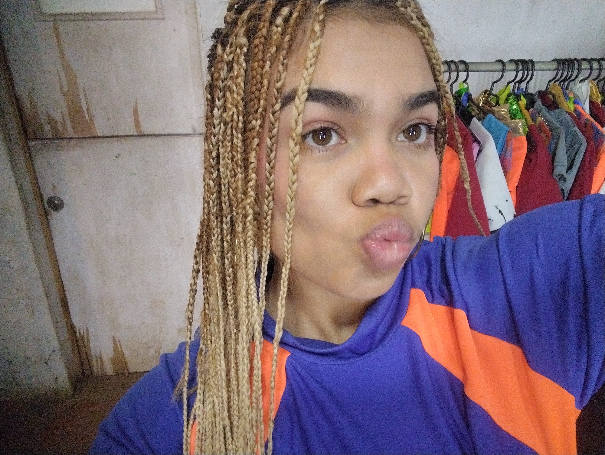 A close-up selfie captures a young person indoors in Porlamar, Venezuela. The individual has light blonde and brown box braids that fall around their face. They have brown eyes and are looking directly at the camera with a playful expression, pursing their glossy, light pink lips. The person is wearing a long-sleeved top, primarily royal blue, with striking orange panels on the shoulders and sleeves.
The background on the left features a light-colored wall or door, possibly white or beige, with visible vertical streaky marks, suggesting wear or texture, and a faint horizontal line. A doorknob is partially visible on the far left. To the right, behind the person's shoulder, a metal clothes rack is laden with various colorful garments on black hangers, including items in red, orange, blue, green, white, and gray. The setting appears to be a casual, personal indoor space, such as a bedroom or dressing area. No visible text is present in the image.