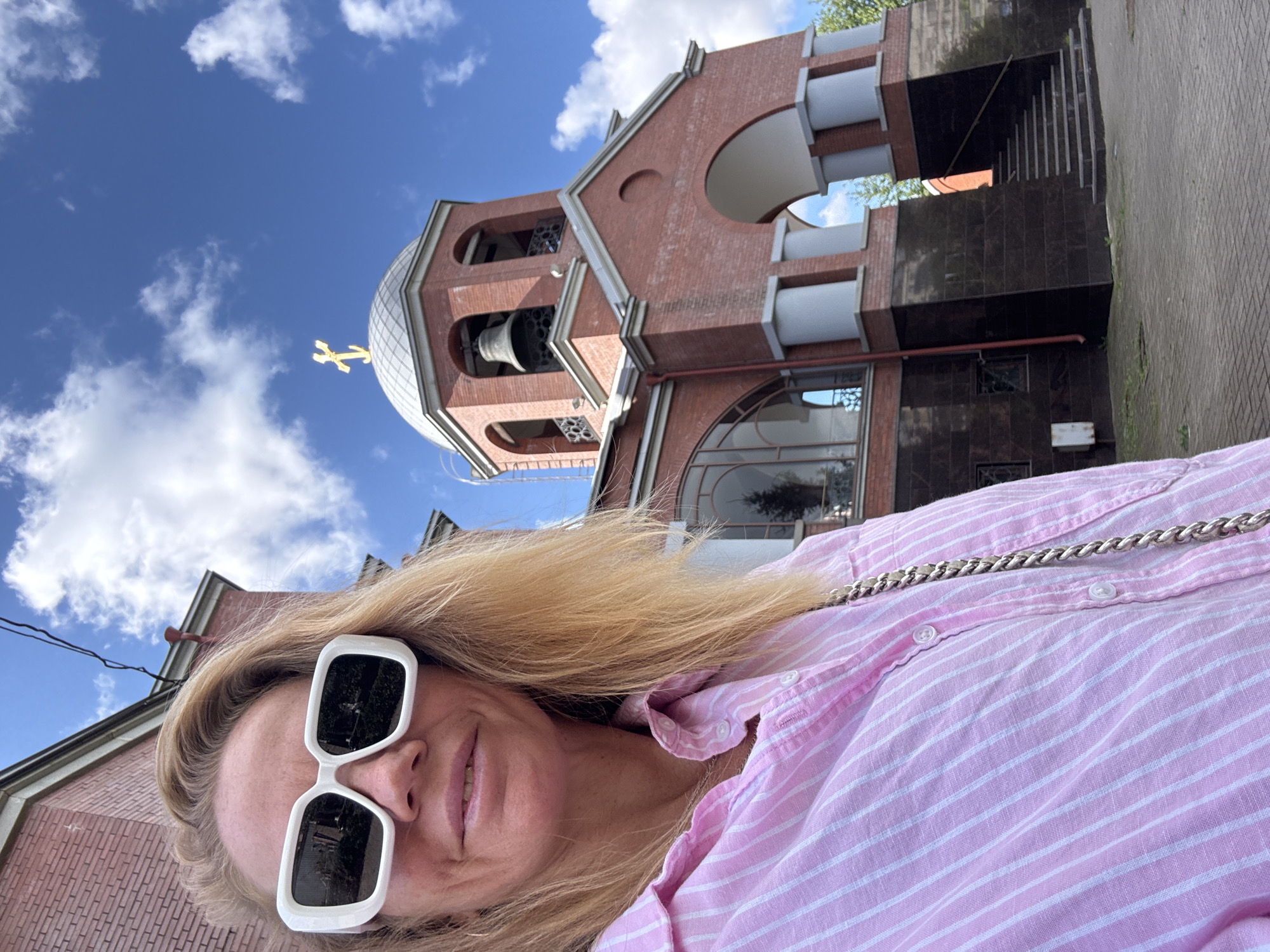A woman wearing large white sunglasses and a pink and white striped button-down shirt takes a selfie in front of a red brick building with a dome and bell tower. The building appears to be an Orthodox church, possibly the Church of the Transfiguration in Saint Petersburg, Russia. The sky is blue with scattered white clouds. The woman's blonde hair is visible. A silver chain purse is slung across her body. The ground in front of the church is paved with bricks.