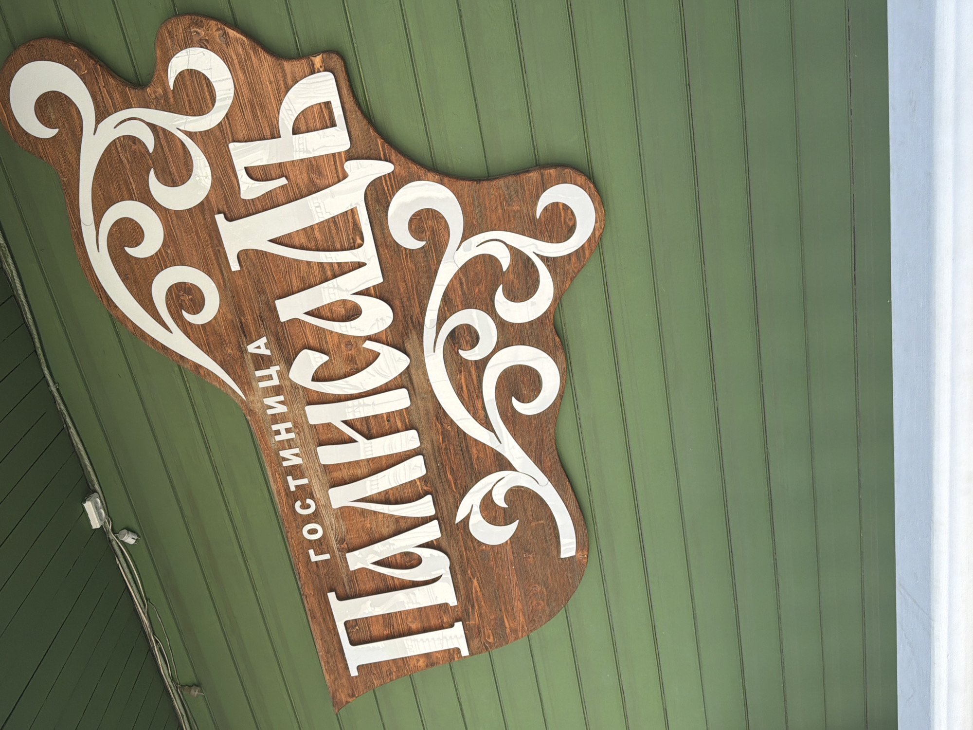 This image shows a wooden sign with white lettering mounted on a green wooden wall. The sign features stylized white decorative elements and the Russian text "ГОСТИНИЦА ПАЛЬСАДЬ," which translates to "Hotel Palisades." The sign is positioned at an angle, with its left side higher than its right. The green wall is composed of horizontal wooden planks, and a white ledge or windowsill is visible at the bottom of the image. Overhead, electrical wiring and a junction box are attached to the green ceiling, which also appears to be made of wooden planks. The overall scene suggests the exterior of a building. This is the entrance sign for the "Palisade" hotel located at 45 Lenina Street in Vologda, Russia.
