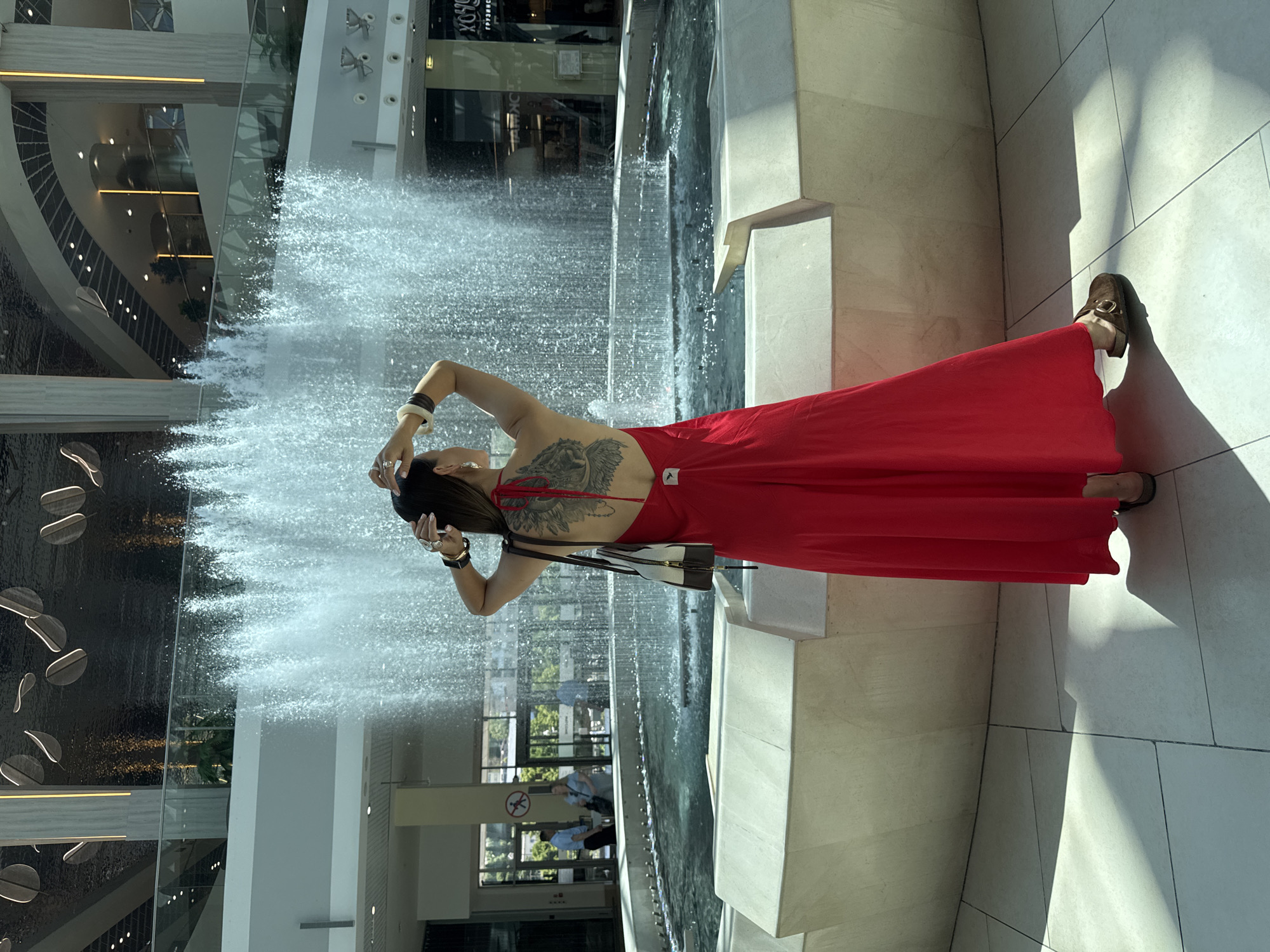 A woman with a large back tattoo stands with her hands on her head in front of an indoor fountain. She is wearing a long red dress, brown slip-on shoes, and several bracelets. The fountain is surrounded by a light-colored stone structure and the floor is tiled. In the background, there are shops and balconies. The overall scene appears to be within a large, modern indoor space, possibly a shopping mall. The image is set in Sochi, Russia.