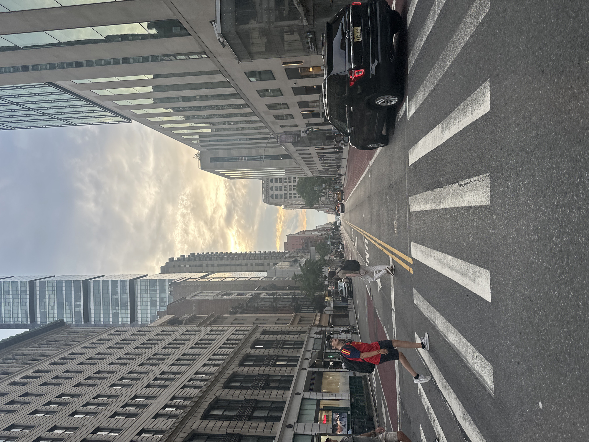 The image depicts a street scene in New York City, United States of America, characterized by tall buildings lining both sides of a wide avenue. In the foreground, a crosswalk with prominent white stripes spans the asphalt road, with a red-painted section indicating a pedestrian zone. Two individuals are crossing the street. On the left, a young person wearing a red and yellow shirt, dark shorts, and white sneakers, with a black backpack, is walking across the crosswalk. To the right, another individual in light-colored pants and white sneakers is also traversing the crosswalk.
A black SUV is positioned in the right lane, stopped at the crosswalk, with its brake lights illuminated. Further down the avenue, other vehicles, including cars and taxis, are visible, suggesting moderate traffic. The buildings are a mix of architectural styles; on the left, a building with many windows and detailed stonework stands, while on the right, a more modern glass-fronted structure rises high. The sky above is partially cloudy, with the sun breaking through, casting a soft light on the scene. The overall impression is of a typical urban environment during daylight hours.