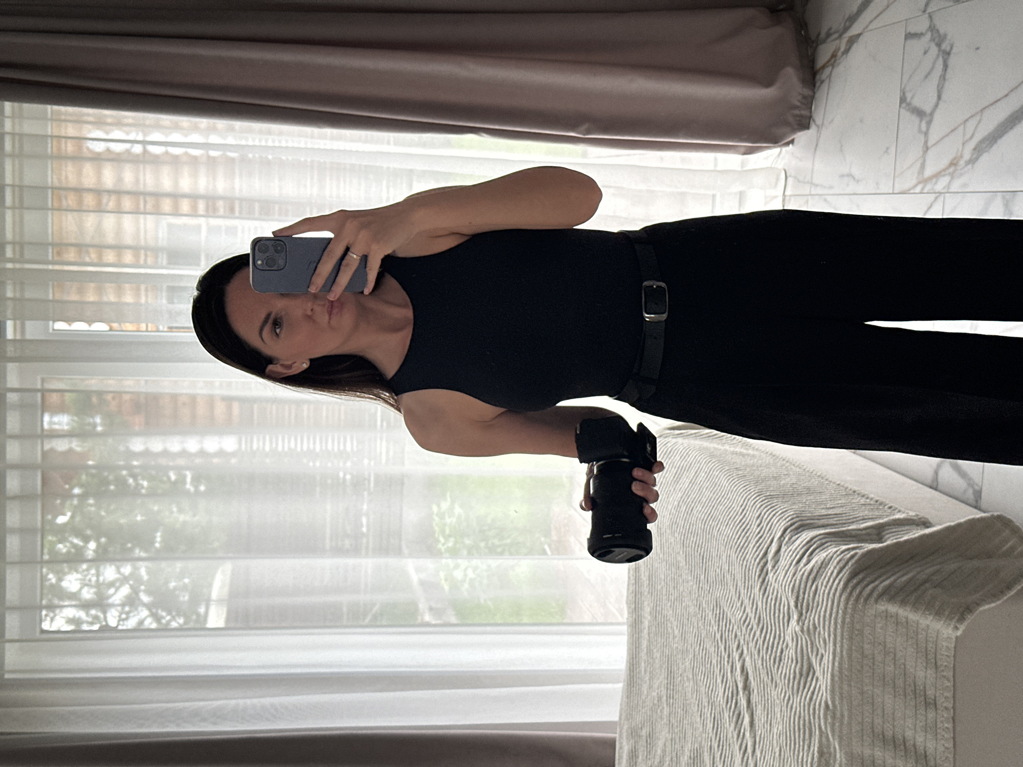 An indoor mirror selfie features a woman posing while holding a smartphone in her right hand and a camera in her left. She has dark hair, is wearing a black sleeveless top, and black pants with a visible belt and silver buckle. A ring is visible on her right hand.
The setting appears to be a modern room, possibly a bedroom, characterized by large windows covered with white blinds and light-colored curtains drawn to the sides. Through the blinds, glimpses of green foliage and a building outline suggest a daytime setting and an outdoor view. To the right of the woman, a bed or sofa is partially visible, covered with a textured, light-colored blanket or throw with a ribbed pattern. Behind this furniture and the woman's right side, there's a light-colored surface with a grey, marble-like pattern, possibly a wall or flooring. The overall lighting is soft, coming primarily from the window. No visible text is present in the image. The scene is set in Chaikovsky, Russia.