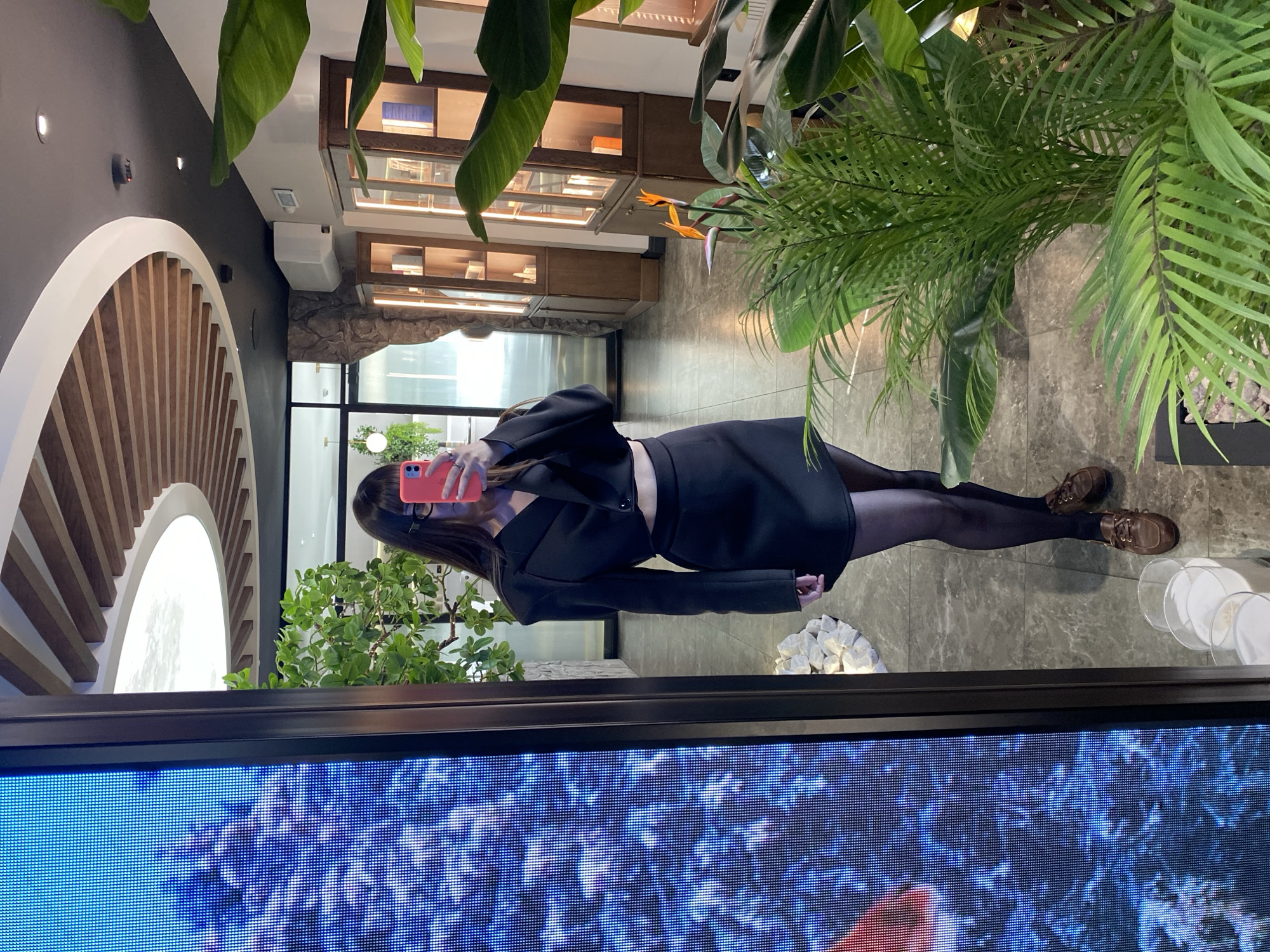 A woman in a black blazer, skirt, and dark tights is reflected in a television screen, holding up a pink phone to take a picture. Her reflection shows she is in a commercial space with display shelves containing merchandise behind her. Artificial greenery and natural light fill the space, and decorative elements like stones and a glass sculpture are visible. Based on the surrounding architecture and information available, it is plausible that this image was taken in a commercial space within the city of Votkinsk, Russia.