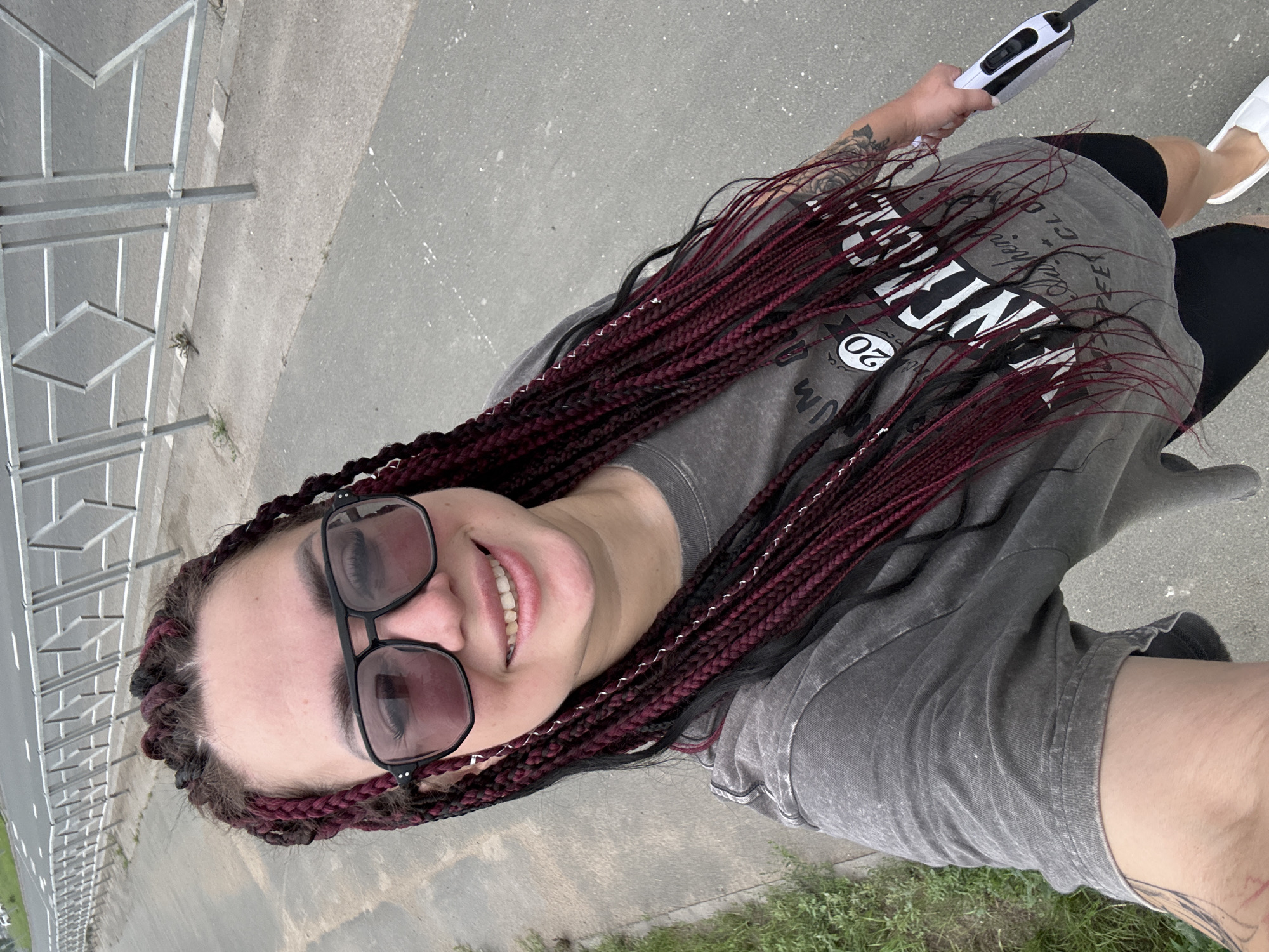 A woman with long, burgundy and black braided hair, wearing oversized black-rimmed glasses and a grey t-shirt with a graphic print, is standing on a paved sidewalk. She is holding a white and black object, possibly a dog leash handle, in her right hand. The background shows a metal fence, a road, and some green grass. The image appears to be taken in Tolyatti, Russia.