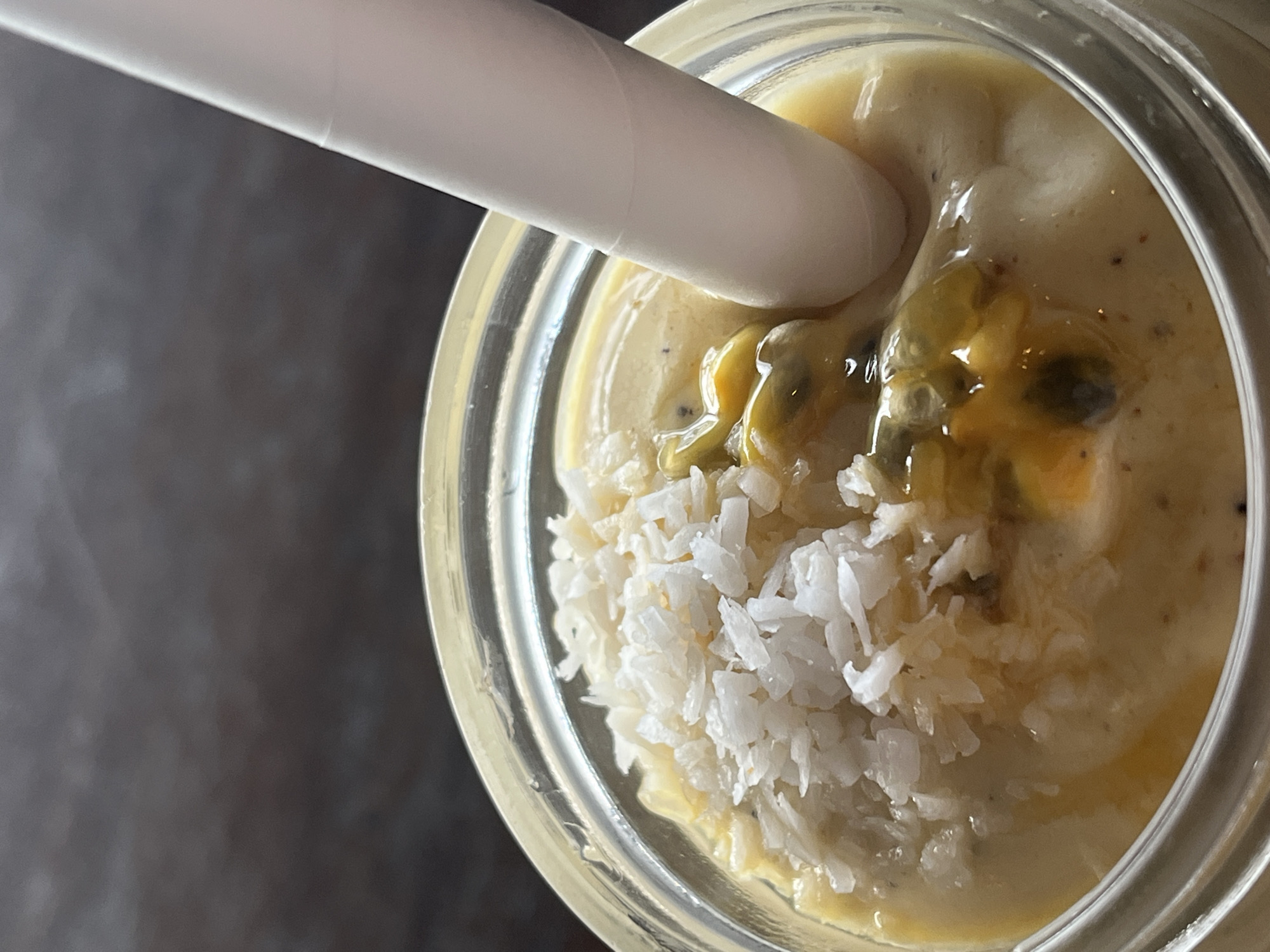A close-up, overhead shot shows a clear glass jar filled with a creamy yellow smoothie. A white paper straw is partially submerged in the smoothie, angled from the upper left towards the center of the jar. The smoothie has a thick, textured consistency, with visible flecks of dark specks, likely seeds or spices, throughout. Scattered on top of the smoothie, particularly on the left side, are generous shavings of white coconut. In the center of the jar, chunks of bright yellow passion fruit pulp and seeds are visible, with some of the gelatinous pulp clinging to the straw. The glass jar has a slight sheen, indicating it is clean and possibly cool. The background is a dark, out-of-focus surface, creating a shallow depth of field that emphasizes the smoothie. The lighting appears to be natural and soft, suggesting an indoor setting during the day. The overall impression is of a refreshing and tropical beverage. Given the context of Baan Chaweng Noi, Thailand, this could be a specialty drink from a local cafe or resort.