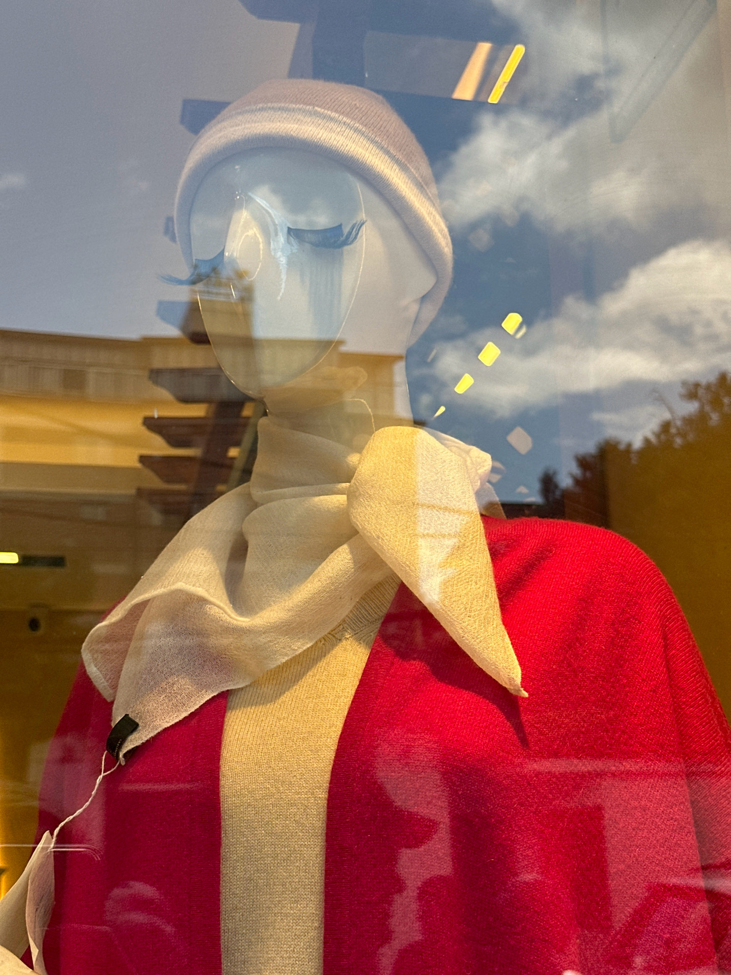 This image shows a mannequin in a store window, likely in Kathmandu Metropolitan City, Nepal. The mannequin is wearing a light-colored beanie, blue false eyelashes, a cream-colored scarf, a light-colored striped sweater, and a red sweater or jacket. The mannequin's face is visible, with prominent eyelashes. The background is a reflection of the sky with clouds, and some yellow rectangular lights are visible, suggesting an indoor setting with reflections from outside. The overall scene is of a retail display, possibly a fashion boutique, in an urban environment.