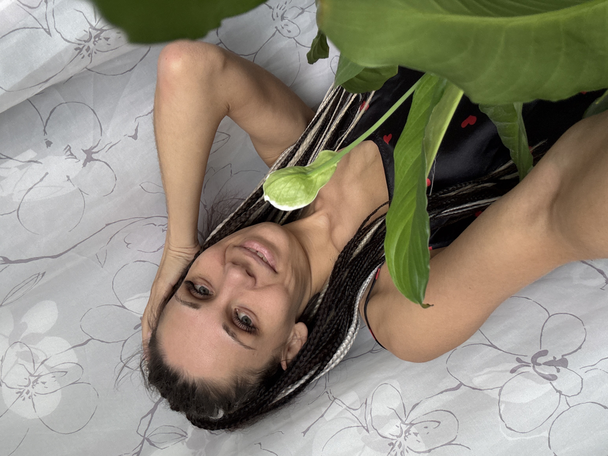 A woman with dark braided hair, featuring some lighter strands, lies on a light-colored fabric patterned with grey floral designs. Her arms are bent, with her hands positioned near her head. She wears a black garment adorned with small red heart patterns. The woman looks directly at the camera with a gentle, calm smile and open eyes. A large green plant with broad leaves and a prominent, spiky light green flower bud partially frames the scene from the foreground and upper right. The setting is indoors, likely a domestic environment, with natural light suggesting daytime. The overall impression is one of relaxation and peace. No text is visible in the image. The scene is situated in Rusanovo, Russia.